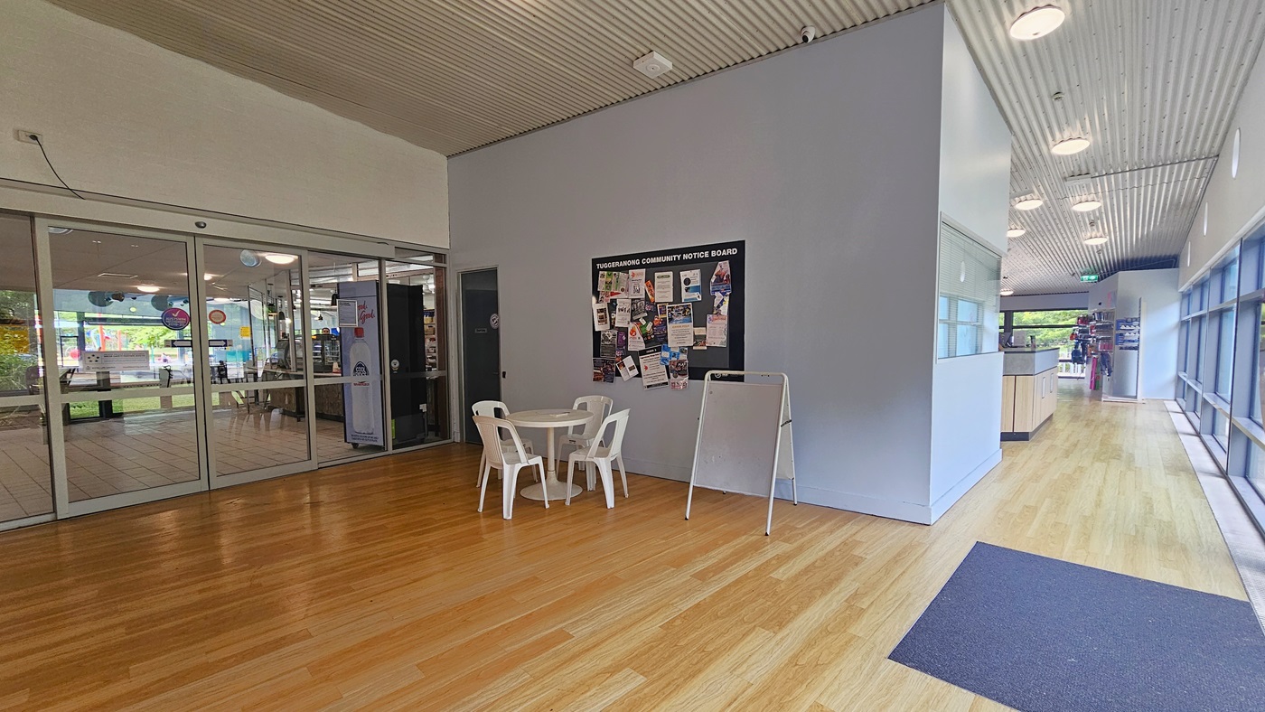 A spacious indoor area with wooden flooring and a corrugated ceiling. On the left, there are glass sliding doors leading to another room with visible decorations. In the centre, a small round table with three white plastic chairs is placed against a wall featuring a community notice board filled with various flyers and notices. On the right, a counter and a corridor with large windows allowing natural light to enter. Ceiling lights are evenly spaced along the corridor. A spacious indoor area with wooden flooring and a corrugated ceiling. On the left, there are glass sliding doors leading to another room with visible decorations. In the centre, a small round table with three white plastic chairs is placed against a wall featuring a community notice board filled with various flyers and notices. On the right, a counter and a corridor with large windows allowing natural light to enter. Ceiling lights are evenly spaced along the corridor.