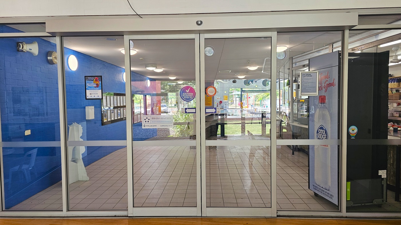 Glass sliding doors with a metal frame, leading into a building. The left side has a blue wall with framed notices and a white dispenser. The right side shows part of a vending machine with a "Cool Ridge" label. Inside, the floor is tiled, and there are several circular ceiling lights. Through the glass, tables and chairs are visible, along with more notices and a view of greenery outside. Stickers and signs are on the doors. Glass sliding doors with a metal frame, leading into a building. The left side has a blue wall with framed notices and a white dispenser. The right side shows part of a vending machine with a "Cool Ridge" label. Inside, the floor is tiled, and there are several circular ceiling lights. Through the glass, tables and chairs are visible, along with more notices and a view of greenery outside. Stickers and signs are on the doors.