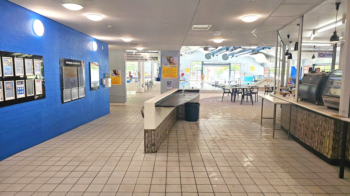 A spacious indoor area with a tiled floor and ceiling lights. On the left, a bright blue wall displays various notices and information boards. In the centre, a long counter with a bin underneath leads towards a pool area visible in the background. On the right, a café counter with a glass display case and hanging lights is adjacent to a seating area with black chairs and tables. The pool area in the background features large windows and visible ductwork on the ceiling. A spacious indoor area with a tiled floor and ceiling lights. On the left, a bright blue wall displays various notices and information boards. In the centre, a long counter with a bin underneath leads towards a pool area visible in the background. On the right, a café counter with a glass display case and hanging lights is adjacent to a seating area with black chairs and tables. The pool area in the background features large windows and visible ductwork on the ceiling.