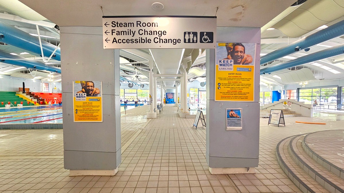 A swimming pool area with tiled flooring and a high ceiling featuring exposed pipes. In the centre, a sign indicates directions to the steam room, family change, and accessible change. On the left, a pool with colourful seating in red, yellow, and green. On the right, a ramp and steps lead to another pool area. Posters with safety information are displayed on the columns. Large windows in the background allow natural light to enter. A swimming pool area with tiled flooring and a high ceiling featuring exposed pipes. In the centre, a sign indicates directions to the steam room, family change, and accessible change. On the left, a pool with colourful seating in red, yellow, and green. On the right, a ramp and steps lead to another pool area. Posters with safety information are displayed on the columns. Large windows in the background allow natural light to enter.