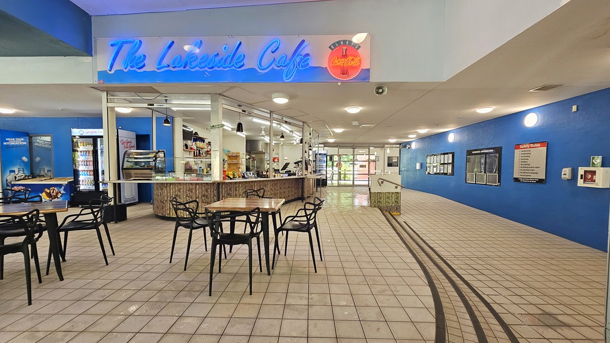 A café with a neon sign reading "The Lakeside Cafe" is on the left. It features a counter with various items and a display case. Several tables and chairs are in front of the counter. The floor is tiled, and the ceiling has recessed lighting. On the right, a blue wall displays information boards and signs. A ramp with a railing leads towards a glass entrance in the background. A café with a neon sign reading "The Lakeside Cafe" is on the left. It features a counter with various items and a display case. Several tables and chairs are in front of the counter. The floor is tiled, and the ceiling has recessed lighting. On the right, a blue wall displays information boards and signs. A ramp with a railing leads towards a glass entrance in the background.
