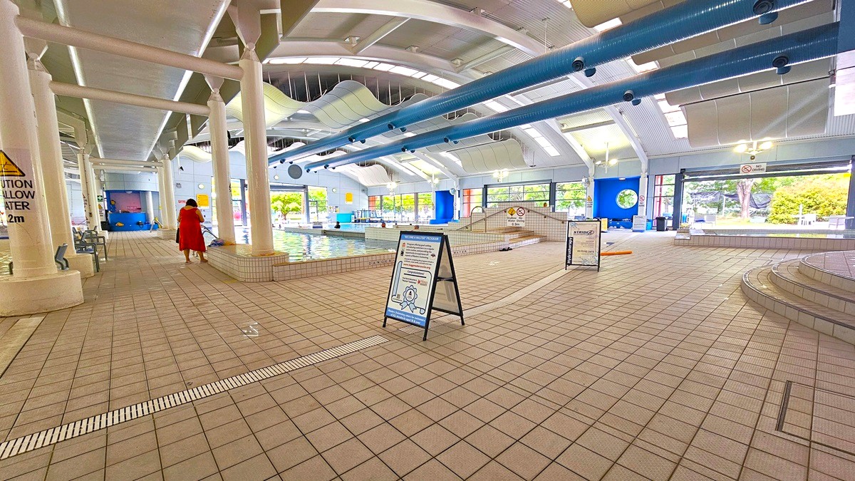 Indoor swimming pool area with tiled flooring and a high ceiling featuring blue pipes. On the left, a person in a red outfit walks towards the pool, which is surrounded by white columns. In the centre, a signboard stands on the floor. The right side has steps leading to another section of the pool. Large windows in the background allow natural light to illuminate the space, and there are caution signs around the pool area. Indoor swimming pool area with tiled flooring and a high ceiling featuring blue pipes. On the left, a person in a red outfit walks towards the pool, which is surrounded by white columns. In the centre, a signboard stands on the floor. The right side has steps leading to another section of the pool. Large windows in the background allow natural light to illuminate the space, and there are caution signs around the pool area.