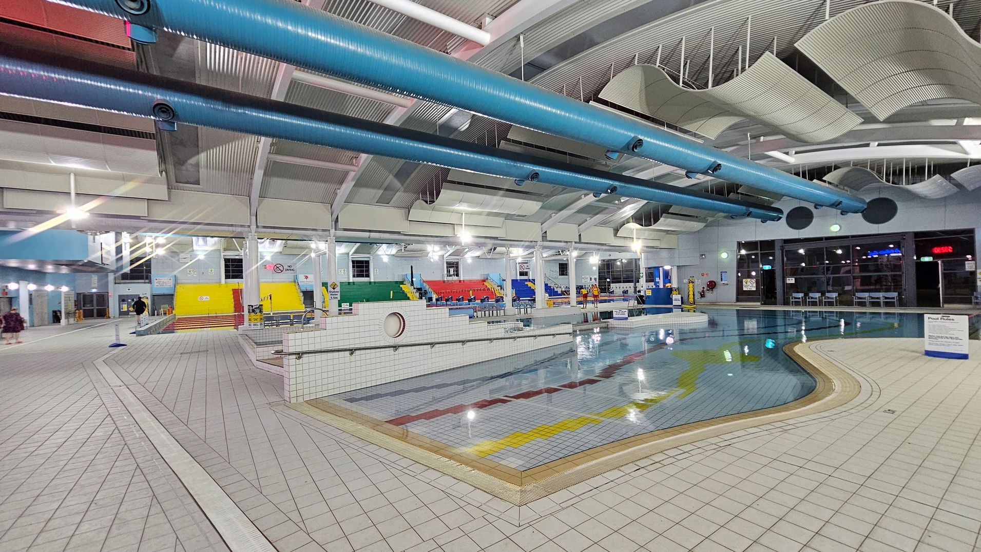 An indoor swimming pool area with a tiled floor and a pool featuring colourful lane markings. On the left, there are steps leading to a yellow wall, with seating in green, red, and blue. The ceiling has large blue pipes and wavy acoustic panels. On the right, large windows and doors are visible, with tables and chairs nearby. The lighting is bright, and the space is spacious and open. An indoor swimming pool area with a tiled floor and a pool featuring colourful lane markings. On the left, there are steps leading to a yellow wall, with seating in green, red, and blue. The ceiling has large blue pipes and wavy acoustic panels. On the right, large windows and doors are visible, with tables and chairs nearby. The lighting is bright, and the space is spacious and open.