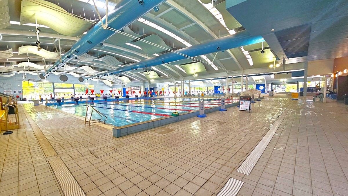 Indoor swimming pool with multiple lanes, marked by red and white flags. The pool is surrounded by a tiled floor. On the left, there are steps leading into the pool. The ceiling features curved, wave-like panels and blue beams. Large windows on the far side allow natural light to enter. The right side has a seating area and informational signs. The space is well-lit with overhead lights. Indoor swimming pool with multiple lanes, marked by red and white flags. The pool is surrounded by a tiled floor. On the left, there are steps leading into the pool. The ceiling features curved, wave-like panels and blue beams. Large windows on the far side allow natural light to enter. The right side has a seating area and informational signs. The space is well-lit with overhead lights.