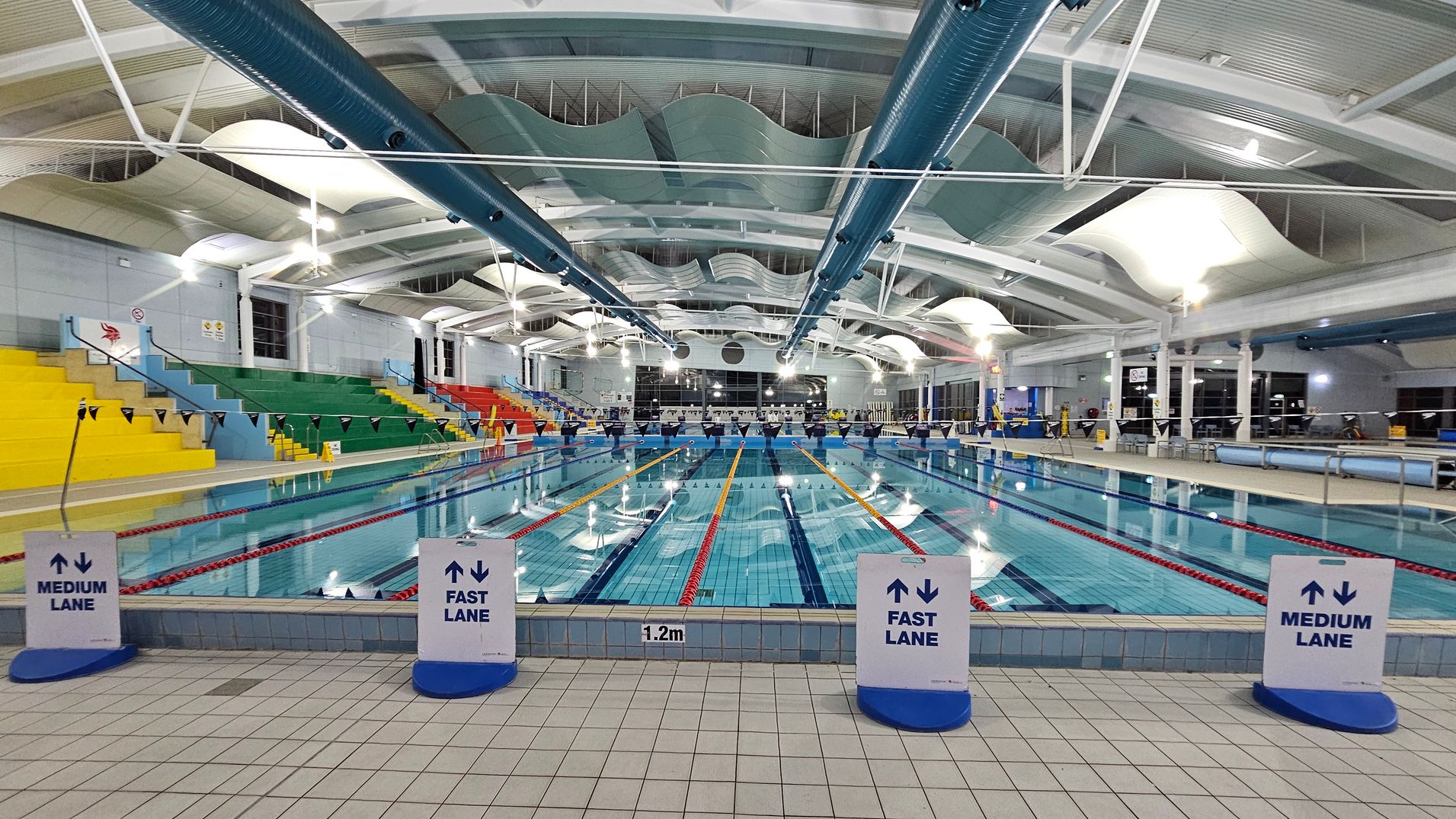 An indoor swimming pool with multiple lanes, each marked by floating lane dividers. Signs at the pool's edge indicate "Medium Lane" and "Fast Lane". On the left, there are tiered seating areas with yellow, green, and red seats. The ceiling features large, curved panels and exposed ductwork. The floor is tiled in light colours, and the pool depth is marked as 1.2 metres. Bright overhead lighting illuminates the space. An indoor swimming pool with multiple lanes, each marked by floating lane dividers. Signs at the pool's edge indicate "Medium Lane" and "Fast Lane". On the left, there are tiered seating areas with yellow, green, and red seats. The ceiling features large, curved panels and exposed ductwork. The floor is tiled in light colours, and the pool depth is marked as 1.2 metres. Bright overhead lighting illuminates the space.