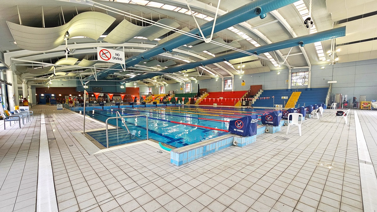 An indoor swimming pool with multiple lanes, surrounded by a tiled deck. On the left, there are chairs and a "No Diving" sign hanging from the ceiling. The right side features more chairs and a few white plastic chairs. In the background, there are colourful spectator stands in red, yellow, and blue. The ceiling has large, curved white panels and exposed beams. The pool area is well-lit with natural light coming through the windows. An indoor swimming pool with multiple lanes, surrounded by a tiled deck. On the left, there are chairs and a "No Diving" sign hanging from the ceiling. The right side features more chairs and a few white plastic chairs. In the background, there are colourful spectator stands in red, yellow, and blue. The ceiling has large, curved white panels and exposed beams. The pool area is well-lit with natural light coming through the windows.