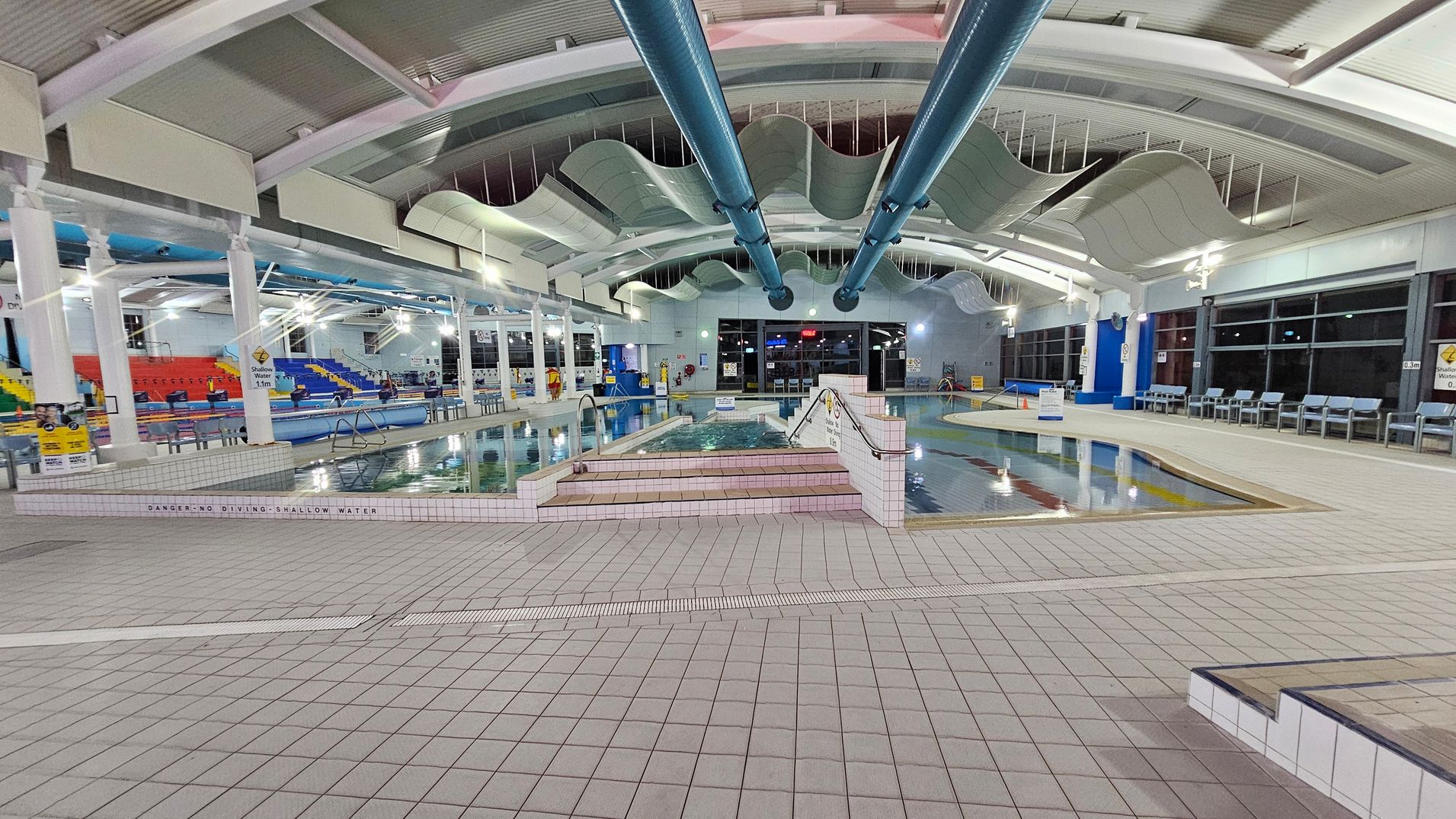 An indoor swimming pool area with a tiled floor and a wavy ceiling design. In the centre, there are two large blue pipes running along the ceiling. The pool is surrounded by a tiled deck, with steps leading into the water. On the left, there are colourful seating areas with red, yellow, and blue seats. On the right, a row of blue chairs lines the wall. Large windows are visible in the background, and various signs are placed around the pool area. An indoor swimming pool area with a tiled floor and a wavy ceiling design. In the centre, there are two large blue pipes running along the ceiling. The pool is surrounded by a tiled deck, with steps leading into the water. On the left, there are colourful seating areas with red, yellow, and blue seats. On the right, a row of blue chairs lines the wall. Large windows are visible in the background, and various signs are placed around the pool area.