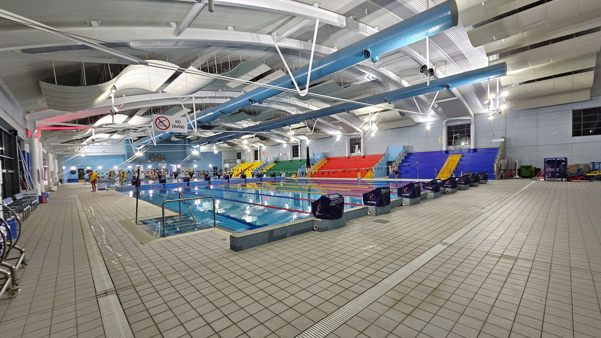 An indoor swimming pool with multiple lanes, surrounded by a tiled floor. On the left, there are chairs and a "No Diving" sign. In the middle, the pool is lined with lane dividers and starting blocks. On the right, there are colourful spectator seats in blue, red, yellow, and green. The ceiling features exposed beams and ductwork, with overhead lighting illuminating the area. An indoor swimming pool with multiple lanes, surrounded by a tiled floor. On the left, there are chairs and a "No Diving" sign. In the middle, the pool is lined with lane dividers and starting blocks. On the right, there are colourful spectator seats in blue, red, yellow, and green. The ceiling features exposed beams and ductwork, with overhead lighting illuminating the area.