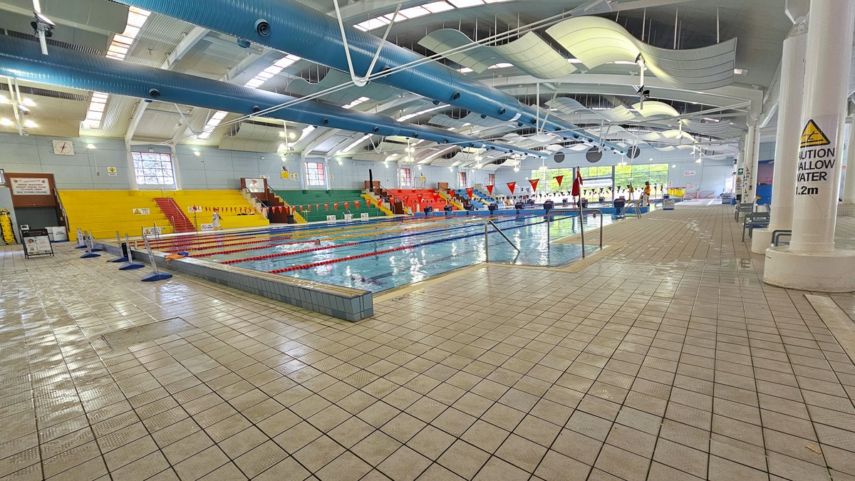 Indoor swimming pool with lanes marked by red and white ropes. On the left, there are yellow and green spectator seats with red stairs. The right side has a tiled pool deck with a caution sign on a pillar. The ceiling features large blue pipes and white curved panels. The floor is covered with light-coloured tiles. Bright overhead lighting and windows provide illumination. Indoor swimming pool with lanes marked by red and white ropes. On the left, there are yellow and green spectator seats with red stairs. The right side has a tiled pool deck with a caution sign on a pillar. The ceiling features large blue pipes and white curved panels. The floor is covered with light-coloured tiles. Bright overhead lighting and windows provide illumination.