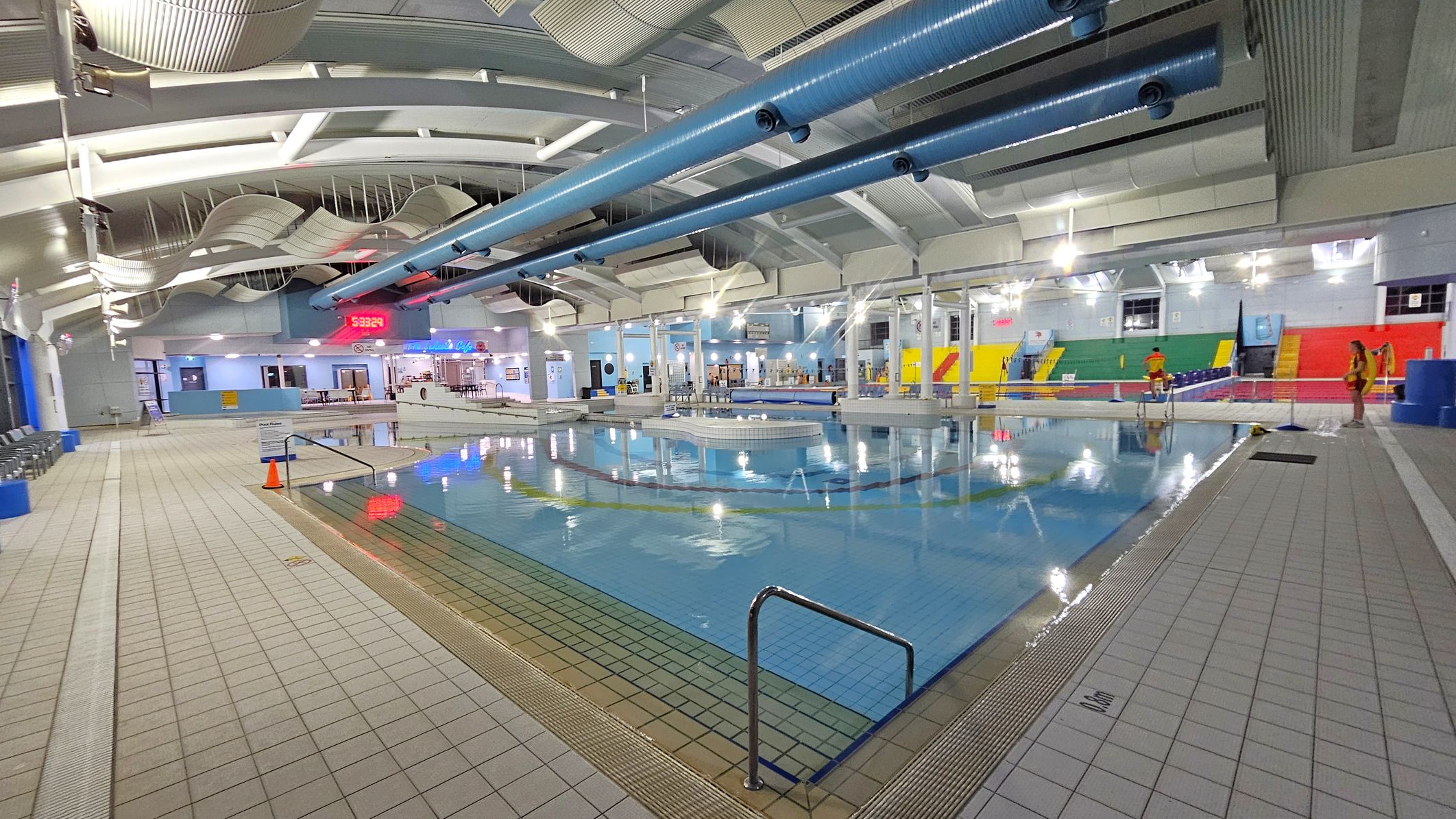 An indoor swimming pool area with a tiled floor surrounding the water. On the left, there are rows of chairs along the wall. The ceiling features curved panels and large blue ventilation ducts. In the centre, a digital clock displays the time. The background includes a café area with tables and chairs. On the right, colourful seating in red, yellow, and green is visible, with a few people standing nearby. The pool has a metal handrail at the edge. An indoor swimming pool area with a tiled floor surrounding the water. On the left, there are rows of chairs along the wall. The ceiling features curved panels and large blue ventilation ducts. In the centre, a digital clock displays the time. The background includes a café area with tables and chairs. On the right, colourful seating in red, yellow, and green is visible, with a few people standing nearby. The pool has a metal handrail at the edge.