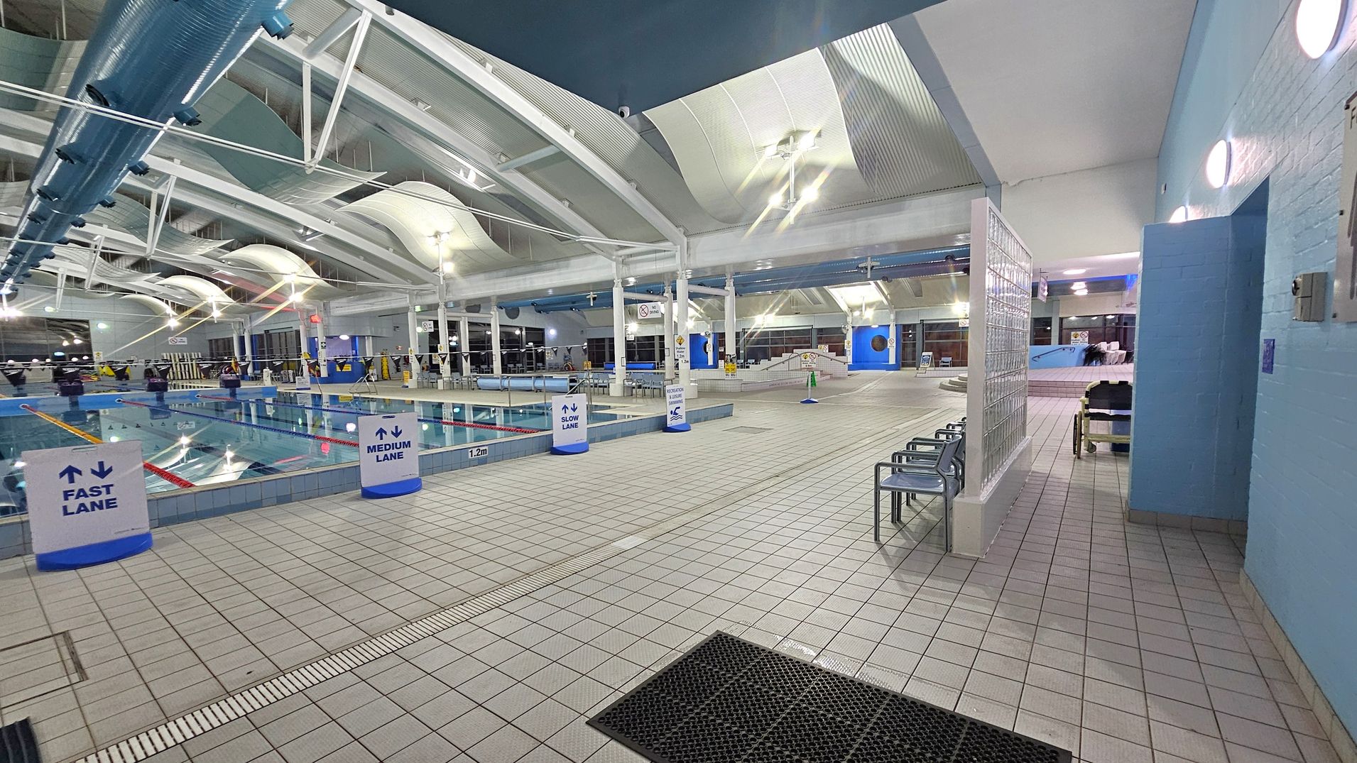 Indoor swimming pool area with lanes marked as fast, medium, and slow. The pool is on the left, with clear water and lane dividers. The floor is tiled in a light colour, with a black mat near the entrance on the right. Several metal chairs are positioned along the wall on the right. The ceiling is high with white beams and large, curved light fixtures. The walls are painted light blue, and there are glass blocks near the entrance. Bright overhead lighting illuminates the space. Indoor swimming pool area with lanes marked as fast, medium, and slow. The pool is on the left, with clear water and lane dividers. The floor is tiled in a light colour, with a black mat near the entrance on the right. Several metal chairs are positioned along the wall on the right. The ceiling is high with white beams and large, curved light fixtures. The walls are painted light blue, and there are glass blocks near the entrance. Bright overhead lighting illuminates the space.
