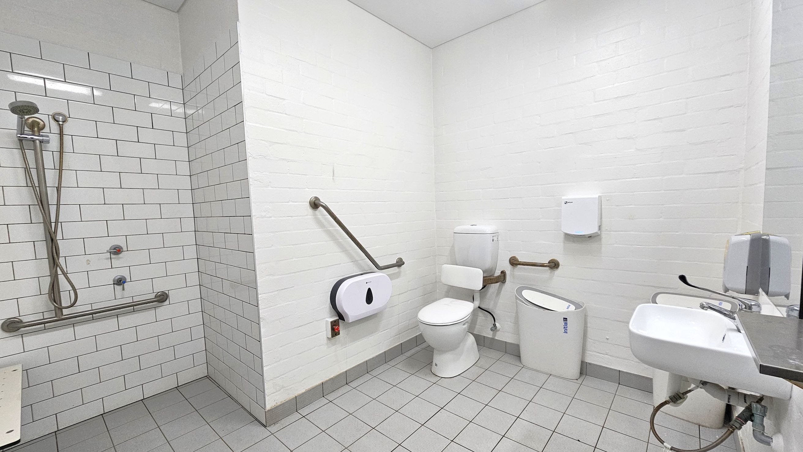 A white-tiled accessible bathroom with a shower area on the left, featuring a handheld showerhead and grab bars. In the centre, there's a toilet with support rails and a sanitary bin nearby. On the right, a small sink with a lever tap is mounted on the wall, accompanied by a soap dispenser. The floor is covered with grey tiles, and the ceiling is plain white. A white-tiled accessible bathroom with a shower area on the left, featuring a handheld showerhead and grab bars. In the centre, there's a toilet with support rails and a sanitary bin nearby. On the right, a small sink with a lever tap is mounted on the wall, accompanied by a soap dispenser. The floor is covered with grey tiles, and the ceiling is plain white.