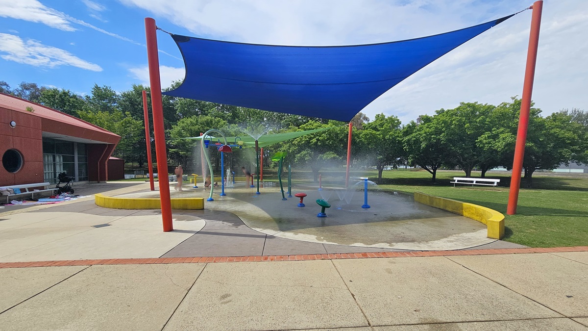 A splash pad with various water features is covered by a large blue shade sail supported by red poles. The ground is a mix of concrete and wet surfaces. On the left, a brick building with circular windows is visible. In the background, there are green trees and a white bench on a grassy area. The sky is partly cloudy. A splash pad with various water features is covered by a large blue shade sail supported by red poles. The ground is a mix of concrete and wet surfaces. On the left, a brick building with circular windows is visible. In the background, there are green trees and a white bench on a grassy area. The sky is partly cloudy.