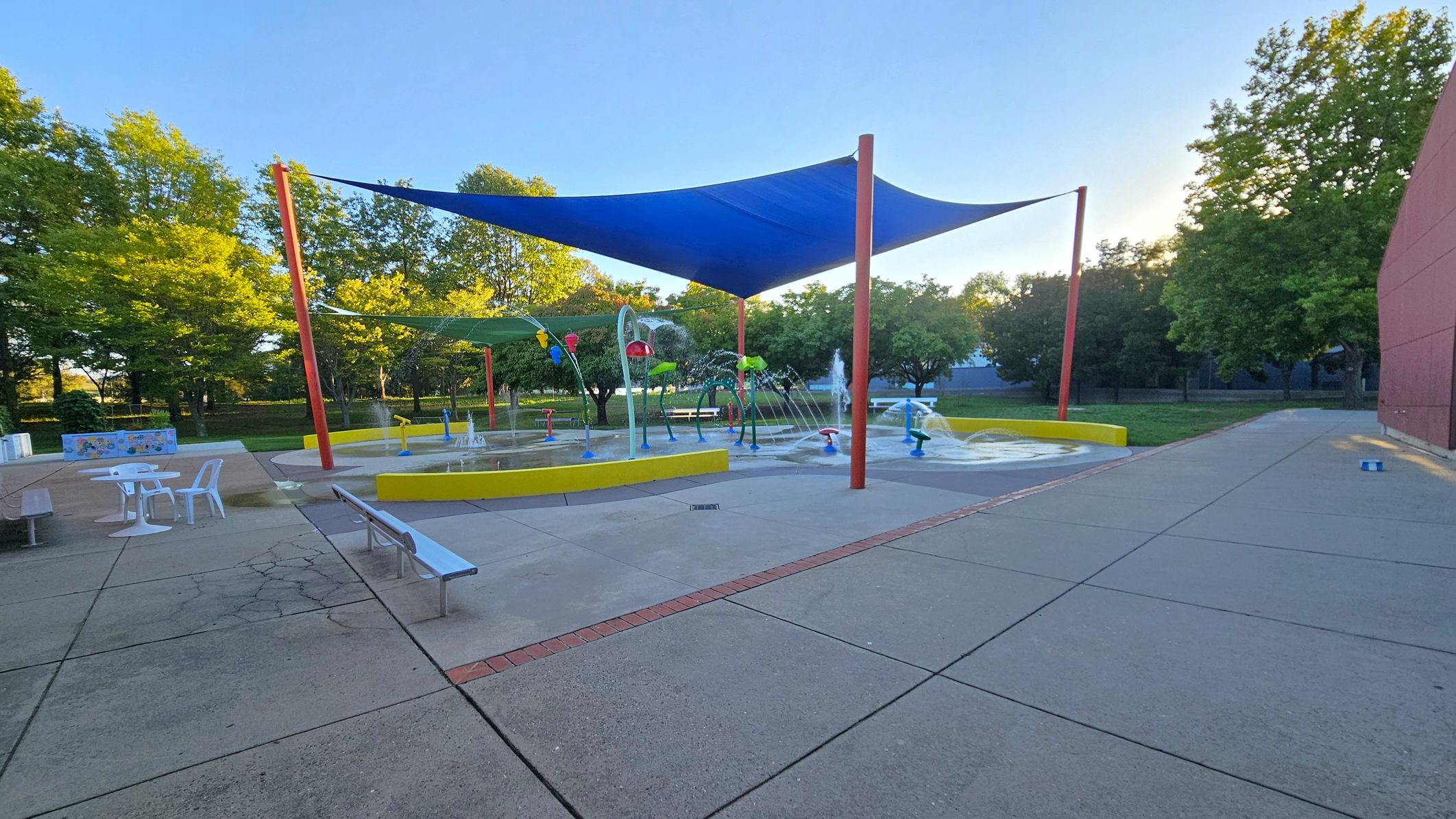 A splash pad with various water features is in the centre, surrounded by a yellow barrier. Blue and green shade sails are supported by red poles above. On the left, there are white tables and chairs on a paved area. The right side has a red brick wall and more pavement. Trees with green foliage are in the background, providing a natural setting. The ground is a mix of concrete and brick patterns. A splash pad with various water features is in the centre, surrounded by a yellow barrier. Blue and green shade sails are supported by red poles above. On the left, there are white tables and chairs on a paved area. The right side has a red brick wall and more pavement. Trees with green foliage are in the background, providing a natural setting. The ground is a mix of concrete and brick patterns.