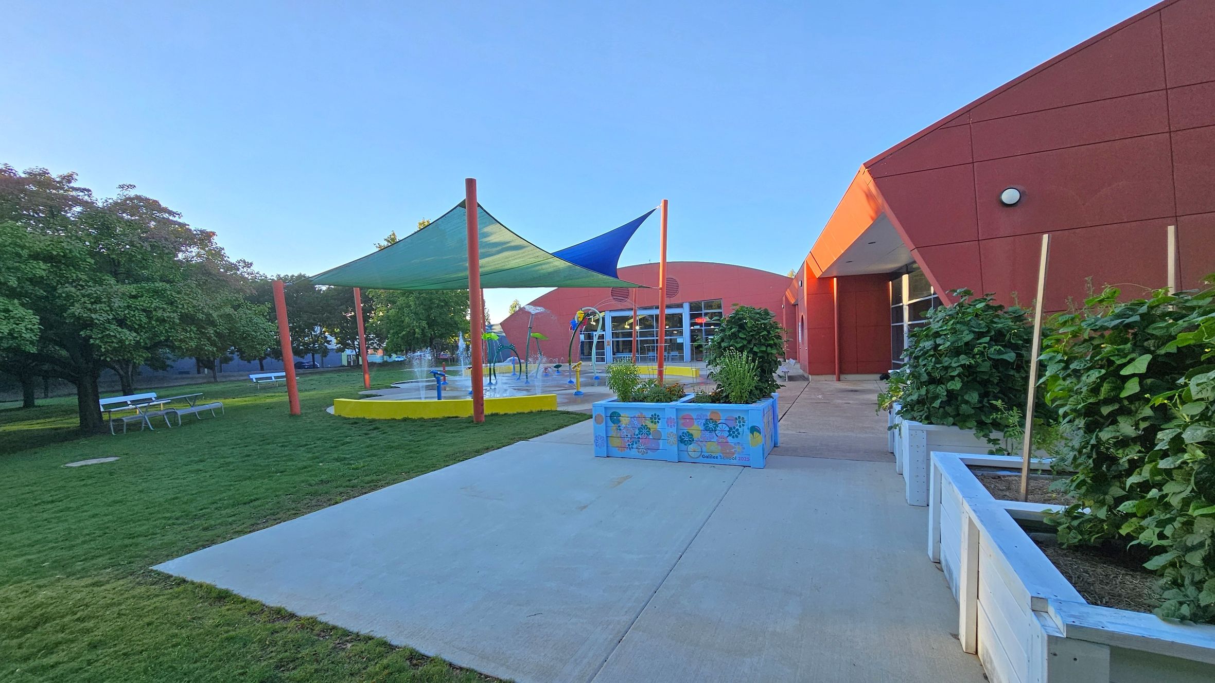 A playground with a splash pad is on the left, featuring a yellow circular base and colourful shade sails. Trees and picnic tables are in the grassy area. On the right, a red building with angular architecture and large windows is visible. In the foreground, there are planter boxes with greenery along a concrete path. A playground with a splash pad is on the left, featuring a yellow circular base and colourful shade sails. Trees and picnic tables are in the grassy area. On the right, a red building with angular architecture and large windows is visible. In the foreground, there are planter boxes with greenery along a concrete path.