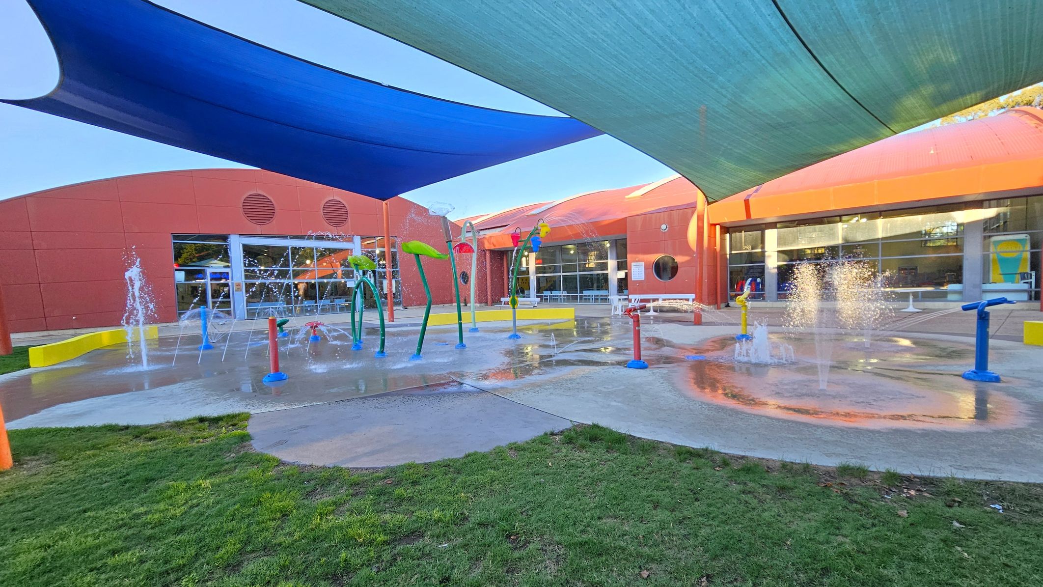 A splash pad with various water features, including fountains and colourful structures, is in the centre. The ground is wet, with some grass visible in the foreground. The background shows a red building with large windows and circular vents. Overhead, blue and green shade sails provide cover. The area is open and designed for recreational water play. A splash pad with various water features, including fountains and colourful structures, is in the centre. The ground is wet, with some grass visible in the foreground. The background shows a red building with large windows and circular vents. Overhead, blue and green shade sails provide cover. The area is open and designed for recreational water play.