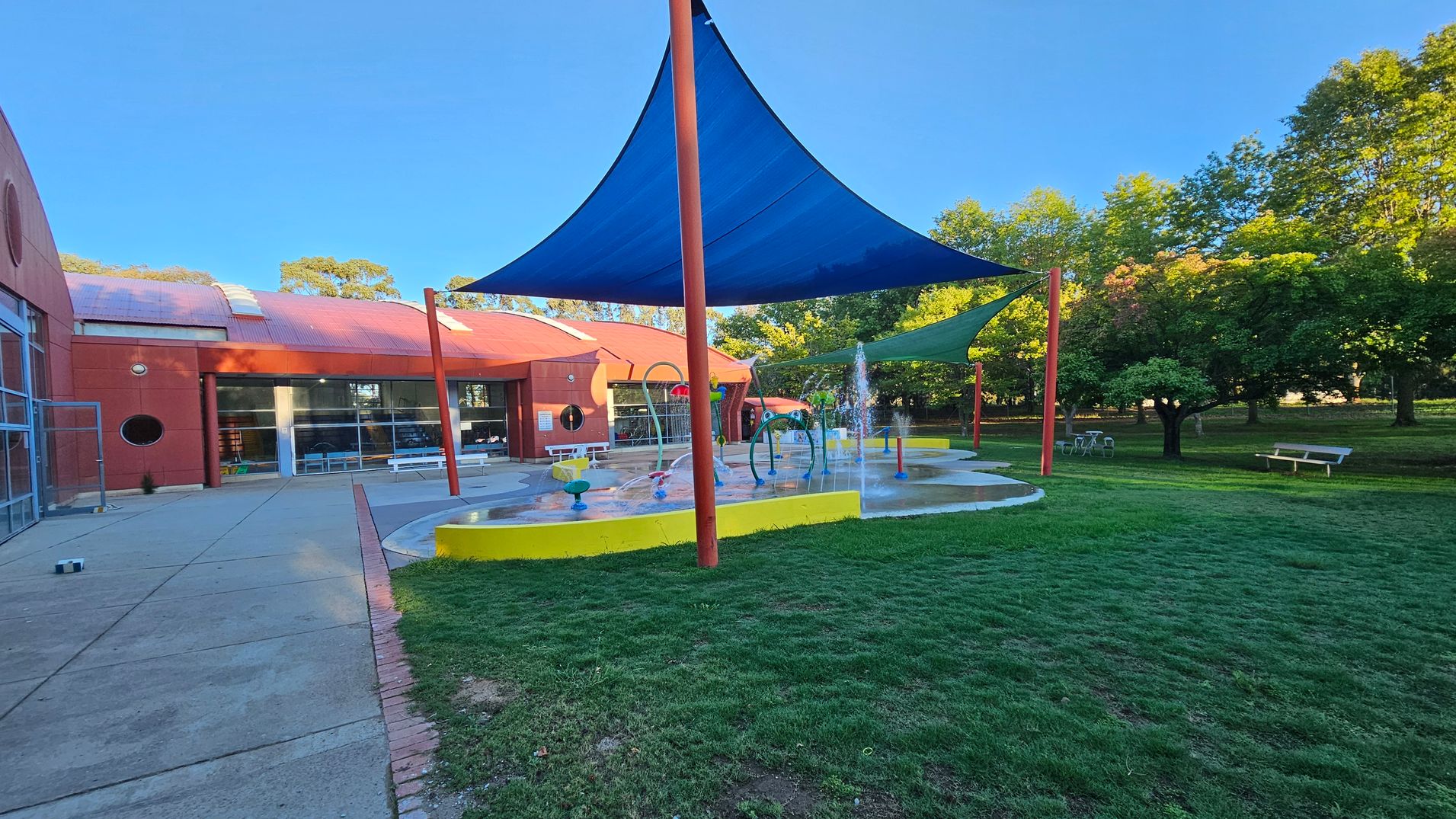 A playground with a splash pad is in the centre, featuring water fountains and colourful play structures. Blue and green shade sails are supported by red poles. On the left, a red brick building with large windows and a circular window is visible. The foreground has a grassy area, while the background includes trees and a bench. The ground is a mix of concrete and grass. A playground with a splash pad is in the centre, featuring water fountains and colourful play structures. Blue and green shade sails are supported by red poles. On the left, a red brick building with large windows and a circular window is visible. The foreground has a grassy area, while the background includes trees and a bench. The ground is a mix of concrete and grass.