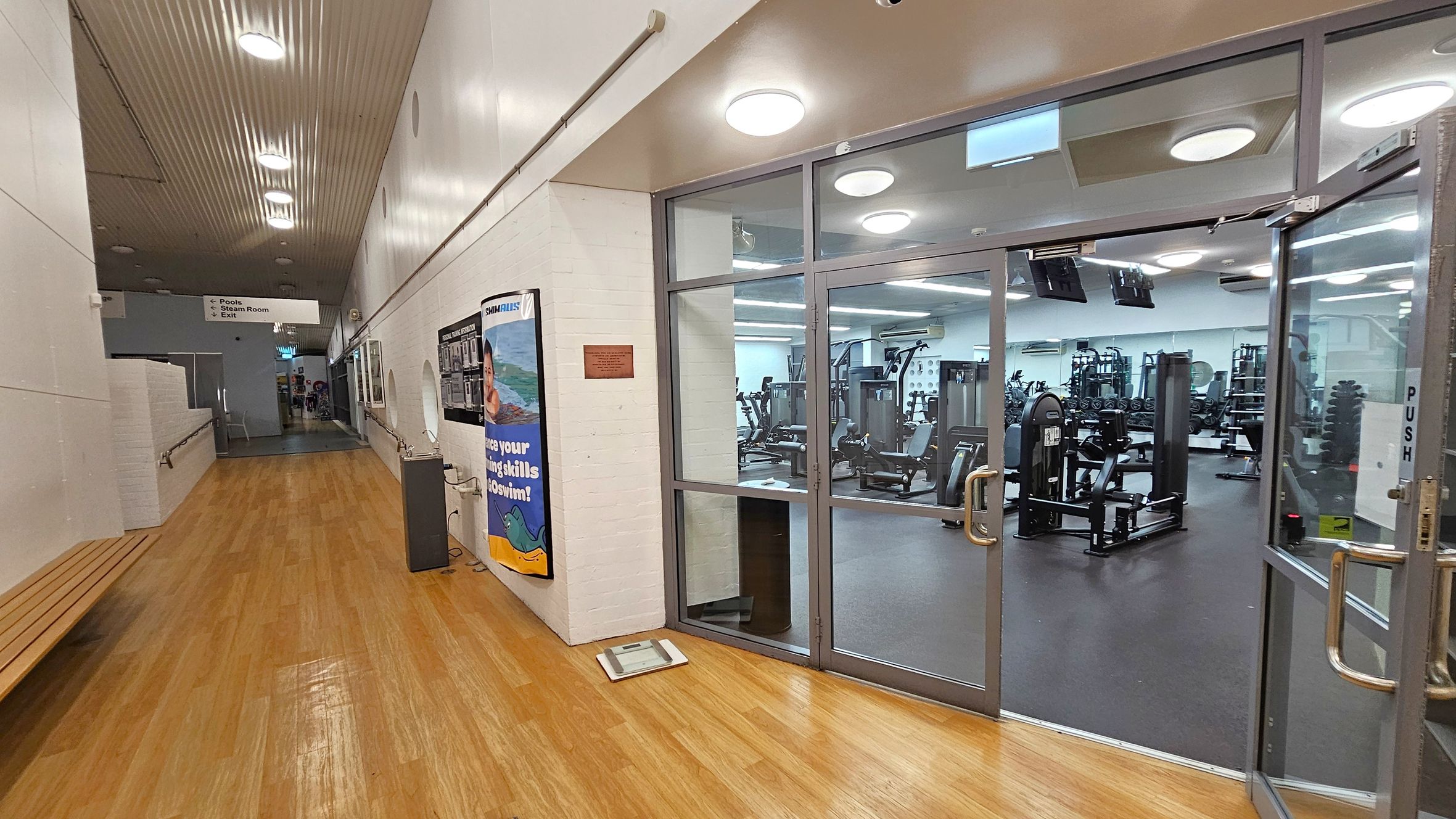 A hallway with a wooden floor and a white ceiling with recessed lights. On the right, there is a glass door leading to a gym with various exercise equipment visible inside. A poster is displayed on the wall next to the door. On the left, there is a wooden bench along the wall. Further down the hallway, a sign indicates directions to pools, a steam room, and an exit. A hallway with a wooden floor and a white ceiling with recessed lights. On the right, there is a glass door leading to a gym with various exercise equipment visible inside. A poster is displayed on the wall next to the door. On the left, there is a wooden bench along the wall. Further down the hallway, a sign indicates directions to pools, a steam room, and an exit.