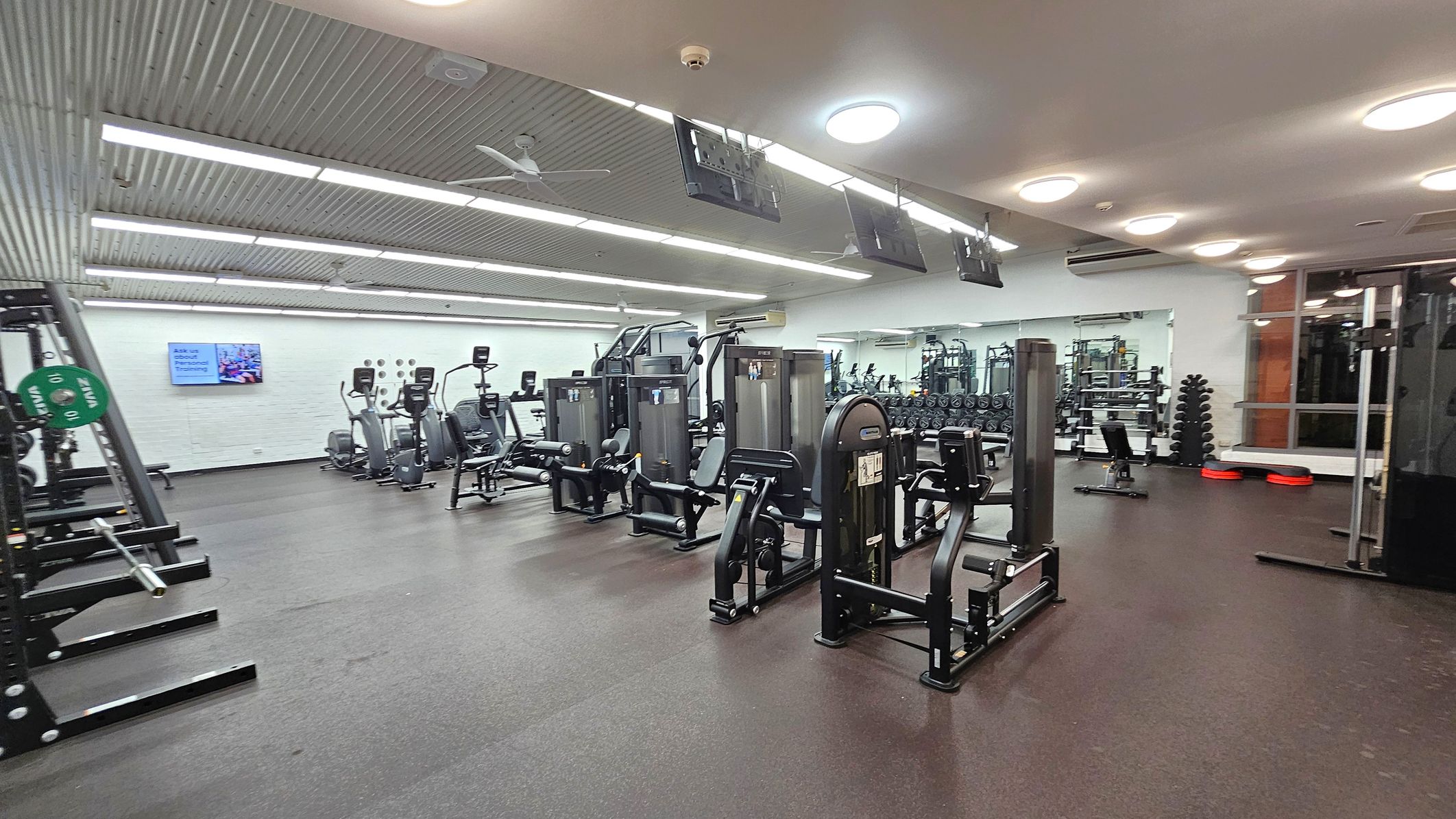 A spacious gym with various exercise machines. On the left, there are weightlifting racks with green weight plates. In the centre, several elliptical machines and strength training equipment are lined up. The right side features a mirrored wall reflecting more gym equipment, including dumbbells and benches. The ceiling has bright lights and a fan, while the floor is covered with dark rubber matting. A wall-mounted screen is visible in the background. A spacious gym with various exercise machines. On the left, there are weightlifting racks with green weight plates. In the centre, several elliptical machines and strength training equipment are lined up. The right side features a mirrored wall reflecting more gym equipment, including dumbbells and benches. The ceiling has bright lights and a fan, while the floor is covered with dark rubber matting. A wall-mounted screen is visible in the background.