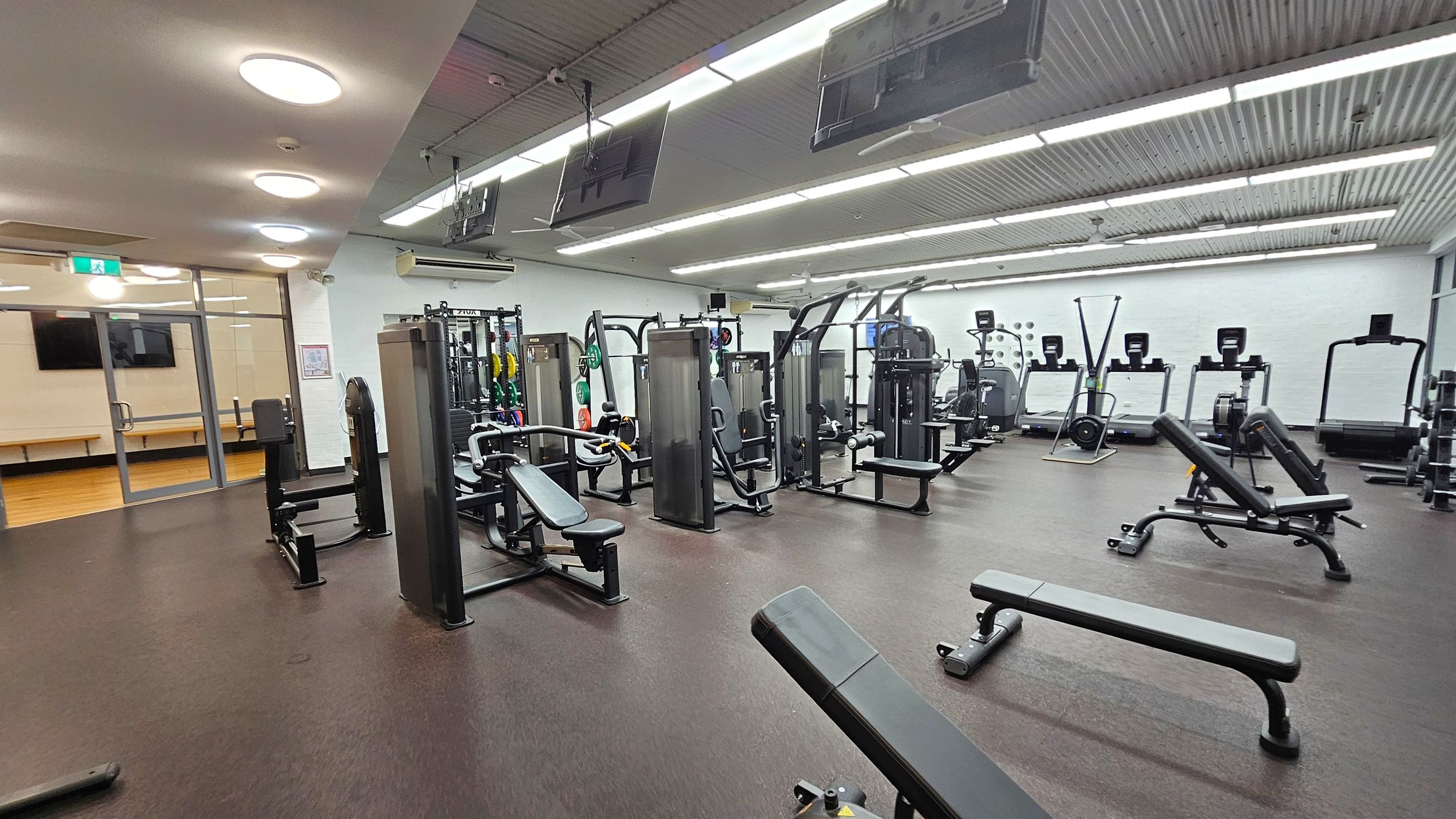 A spacious gym with various exercise equipment. On the left, there are weight machines and benches. In the centre, more weight machines and benches are arranged neatly. On the right, treadmills and rowing machines are lined up against the wall. The floor is dark and rubberised, while the ceiling features bright, recessed lighting. Large windows and a glass door are visible on the far left, leading to another room with wooden flooring. A spacious gym with various exercise equipment. On the left, there are weight machines and benches. In the centre, more weight machines and benches are arranged neatly. On the right, treadmills and rowing machines are lined up against the wall. The floor is dark and rubberised, while the ceiling features bright, recessed lighting. Large windows and a glass door are visible on the far left, leading to another room with wooden flooring.