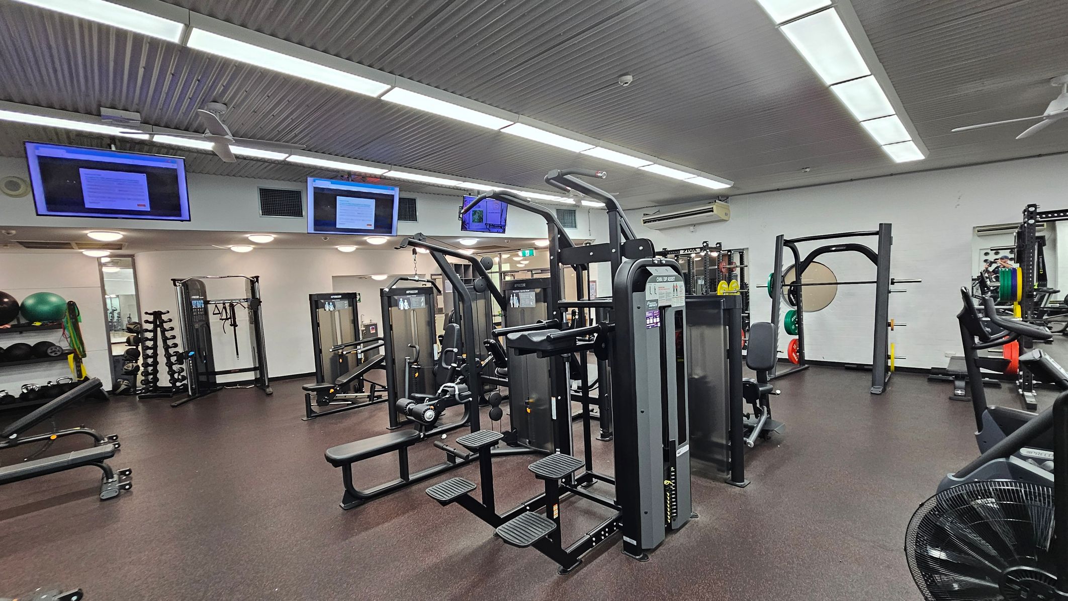 A gym with various exercise equipment. In the centre, there are weight machines and benches. On the left, shelves hold exercise balls and resistance bands, with a rack of dumbbells nearby. On the right, a squat rack with weight plates is visible. The ceiling has fluorescent lights and mounted screens displaying information. The floor is covered with dark rubber matting. A gym with various exercise equipment. In the centre, there are weight machines and benches. On the left, shelves hold exercise balls and resistance bands, with a rack of dumbbells nearby. On the right, a squat rack with weight plates is visible. The ceiling has fluorescent lights and mounted screens displaying information. The floor is covered with dark rubber matting.