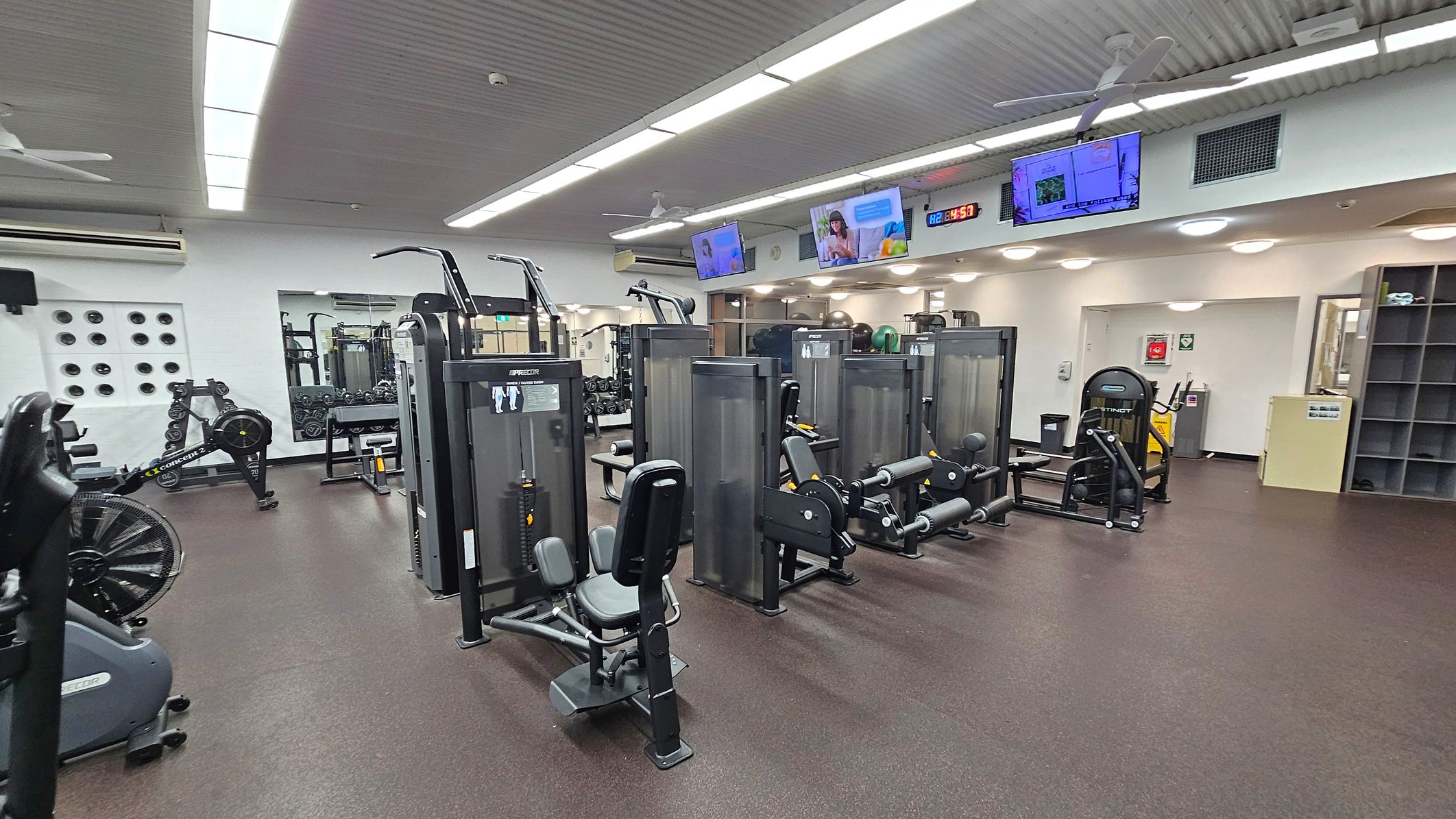 A gym with various exercise machines arranged in rows. On the left, there is a rowing machine and a wall with circular ventilation holes. In the centre, several weight machines are positioned, including leg and arm equipment. On the right, there are more machines and a shelving unit. The floor is dark with a speckled pattern, and the ceiling has fluorescent lights and ceiling fans. Several screens are mounted on the wall displaying different content. A gym with various exercise machines arranged in rows. On the left, there is a rowing machine and a wall with circular ventilation holes. In the centre, several weight machines are positioned, including leg and arm equipment. On the right, there are more machines and a shelving unit. The floor is dark with a speckled pattern, and the ceiling has fluorescent lights and ceiling fans. Several screens are mounted on the wall displaying different content.