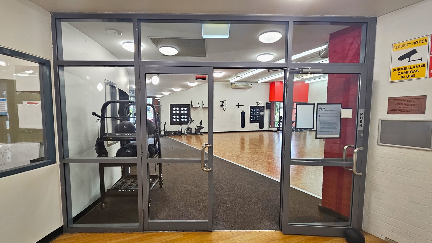 Glass doors with metal frames lead into a gym area. Inside, exercise equipment like ellipticals and punching bags are visible. The floor is wooden, and the ceiling has circular lights. On the right wall, there's a yellow sign indicating surveillance cameras are in use, along with a plaque. The left side shows a partial view of another room through a window. Glass doors with metal frames lead into a gym area. Inside, exercise equipment like ellipticals and punching bags are visible. The floor is wooden, and the ceiling has circular lights. On the right wall, there's a yellow sign indicating surveillance cameras are in use, along with a plaque. The left side shows a partial view of another room through a window.