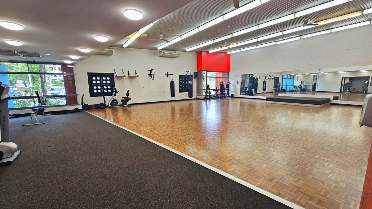 A spacious gym with a wooden floor in the centre and a carpeted area on the left. The left side features exercise equipment, including stationary bikes and a treadmill, near large windows. The right side has a mirrored wall and a raised platform. The ceiling is lined with fluorescent lights, and the walls are adorned with various fitness accessories. A red accent wall is visible in the background. A spacious gym with a wooden floor in the centre and a carpeted area on the left. The left side features exercise equipment, including stationary bikes and a treadmill, near large windows. The right side has a mirrored wall and a raised platform. The ceiling is lined with fluorescent lights, and the walls are adorned with various fitness accessories. A red accent wall is visible in the background.