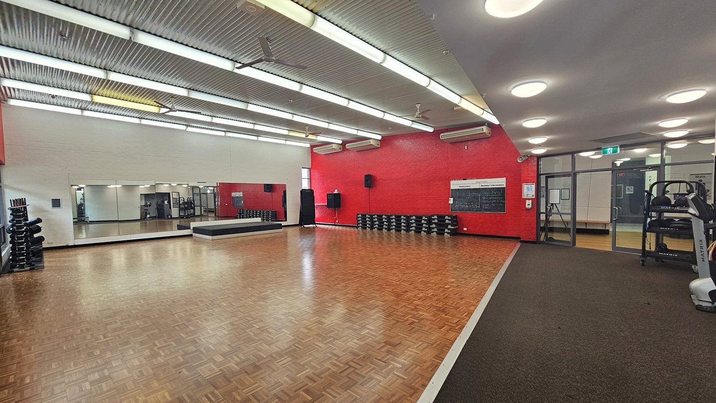 A spacious gym room with a wooden parquet floor. On the left, there are stacks of dumbbells and a large mirror covering the wall. The back wall is painted red and features a row of stacked step platforms and a blackboard. On the right, there is a treadmill and a glass door leading to another room. The ceiling is metallic with fluorescent lights, and the floor transitions to a carpeted area near the treadmill. A spacious gym room with a wooden parquet floor. On the left, there are stacks of dumbbells and a large mirror covering the wall. The back wall is painted red and features a row of stacked step platforms and a blackboard. On the right, there is a treadmill and a glass door leading to another room. The ceiling is metallic with fluorescent lights, and the floor transitions to a carpeted area near the treadmill.