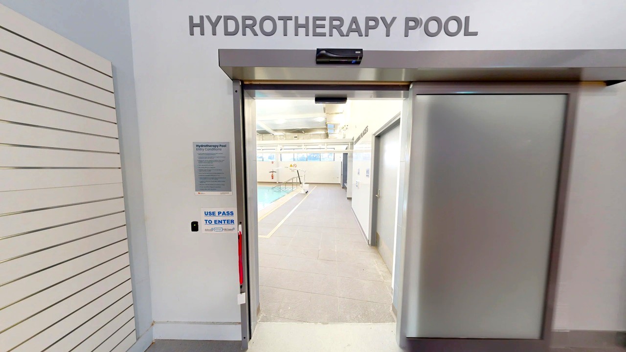 Entrance to a hydrotherapy pool area. The door is metallic with a sign above reading "HYDROTHERAPY POOL". To the left, there is a wall with horizontal lines and a sign instructing to use a pass to enter. Inside, a pool is visible with a pathway leading to it. The ceiling has exposed pipes and lighting fixtures. Entrance to a hydrotherapy pool area. The door is metallic with a sign above reading "HYDROTHERAPY POOL". To the left, there is a wall with horizontal lines and a sign instructing to use a pass to enter. Inside, a pool is visible with a pathway leading to it. The ceiling has exposed pipes and lighting fixtures.