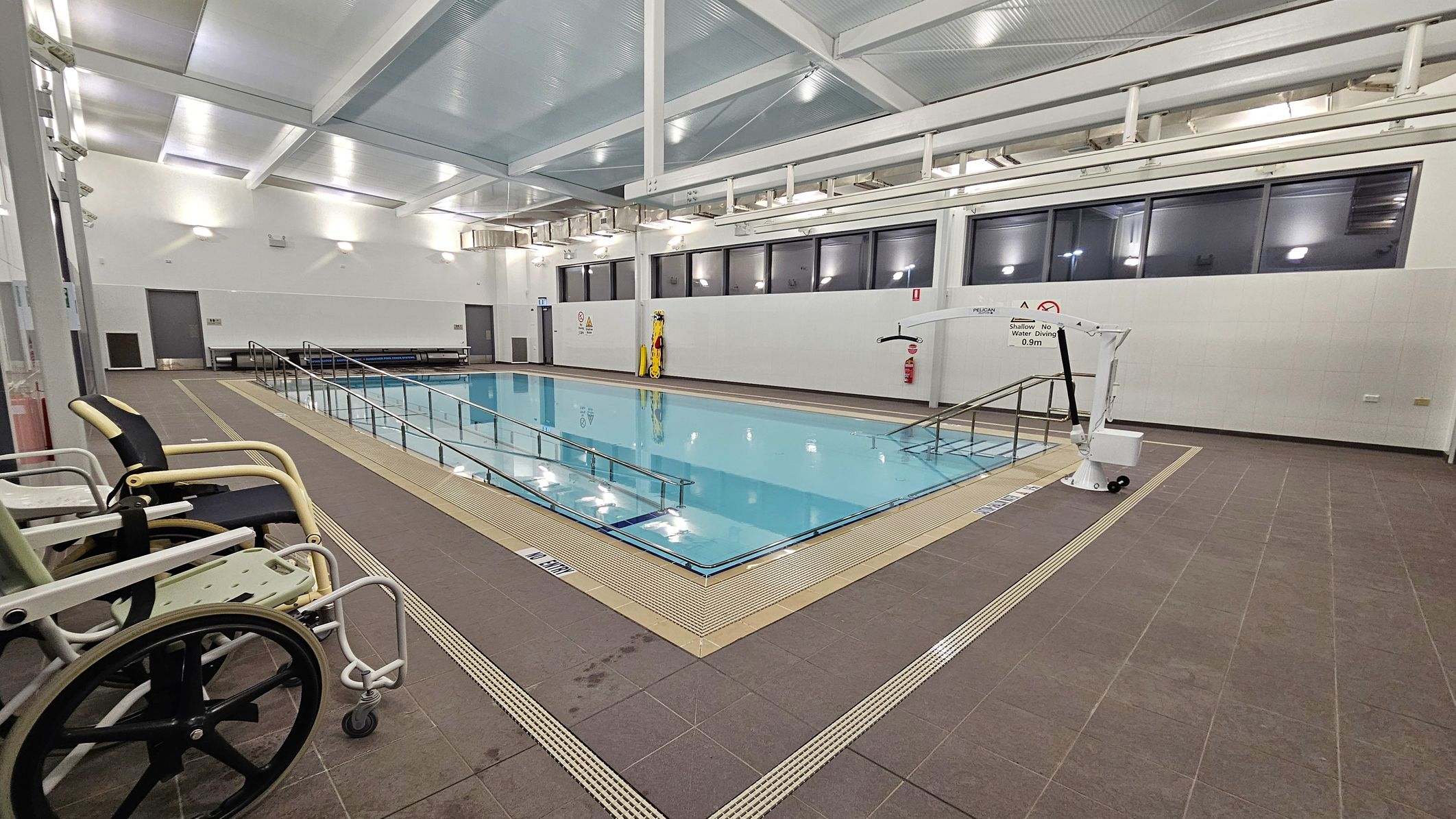 An indoor swimming pool with a ramp leading into the water, surrounded by a brown tiled floor. On the left, there are two wheelchairs positioned near the pool's edge. The right side features a mechanical pool lift. The back wall has large windows and a few doors, with a bench and safety equipment visible. The ceiling is high with exposed beams and bright lighting. An indoor swimming pool with a ramp leading into the water, surrounded by a brown tiled floor. On the left, there are two wheelchairs positioned near the pool's edge. The right side features a mechanical pool lift. The back wall has large windows and a few doors, with a bench and safety equipment visible. The ceiling is high with exposed beams and bright lighting.