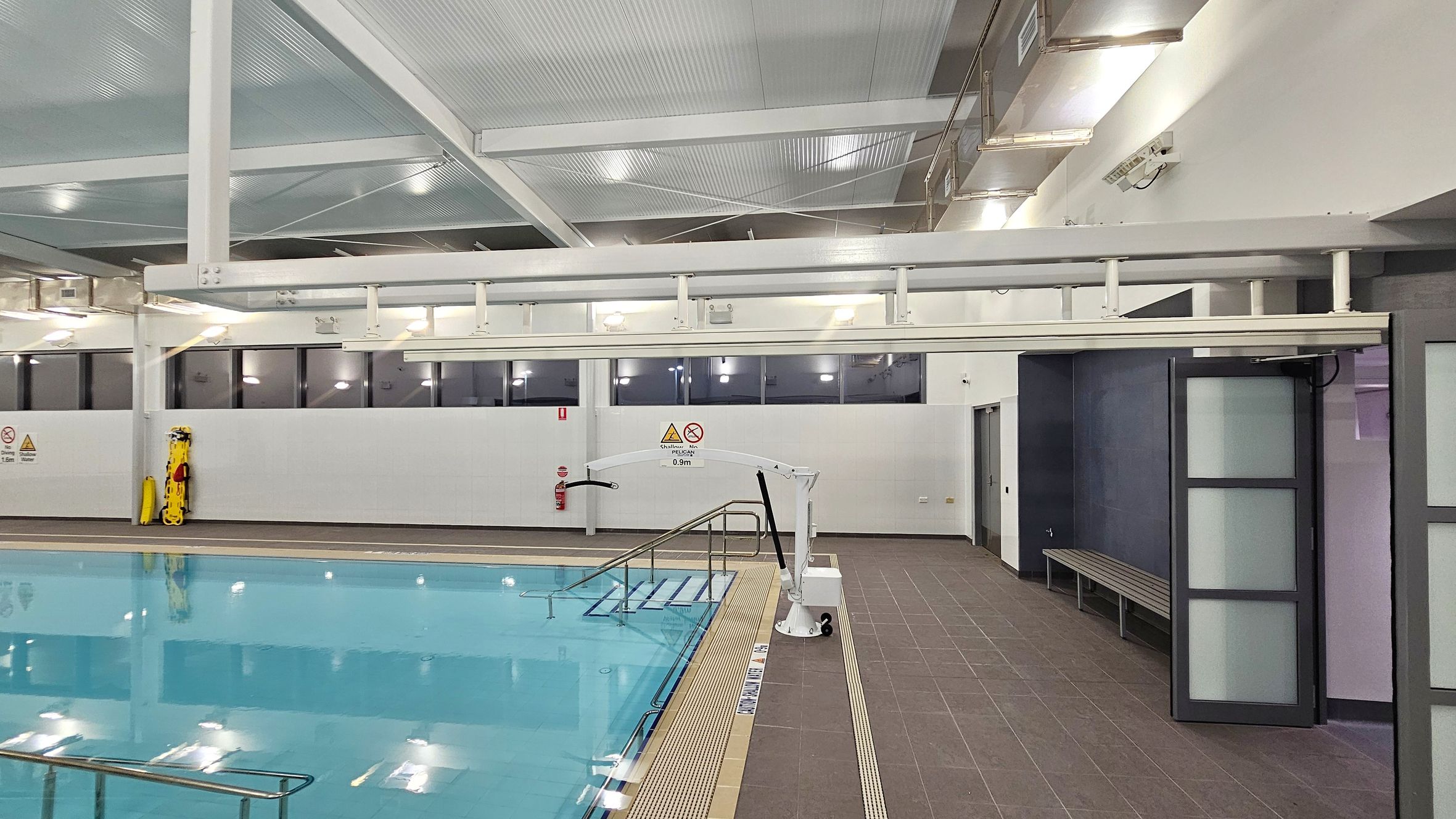 Indoor swimming pool area with a clear blue pool on the left. A metal handrail descends into the water. On the right, a brown tiled floor leads to a wall with a bench and a partially open door with frosted glass panels. Above, a white ceiling with exposed beams and lights. A pool lift is positioned near the pool edge. Windows line the upper wall, and a yellow safety board is mounted on the wall. Warning signs are visible near the pool. Indoor swimming pool area with a clear blue pool on the left. A metal handrail descends into the water. On the right, a brown tiled floor leads to a wall with a bench and a partially open door with frosted glass panels. Above, a white ceiling with exposed beams and lights. A pool lift is positioned near the pool edge. Windows line the upper wall, and a yellow safety board is mounted on the wall. Warning signs are visible near the pool.