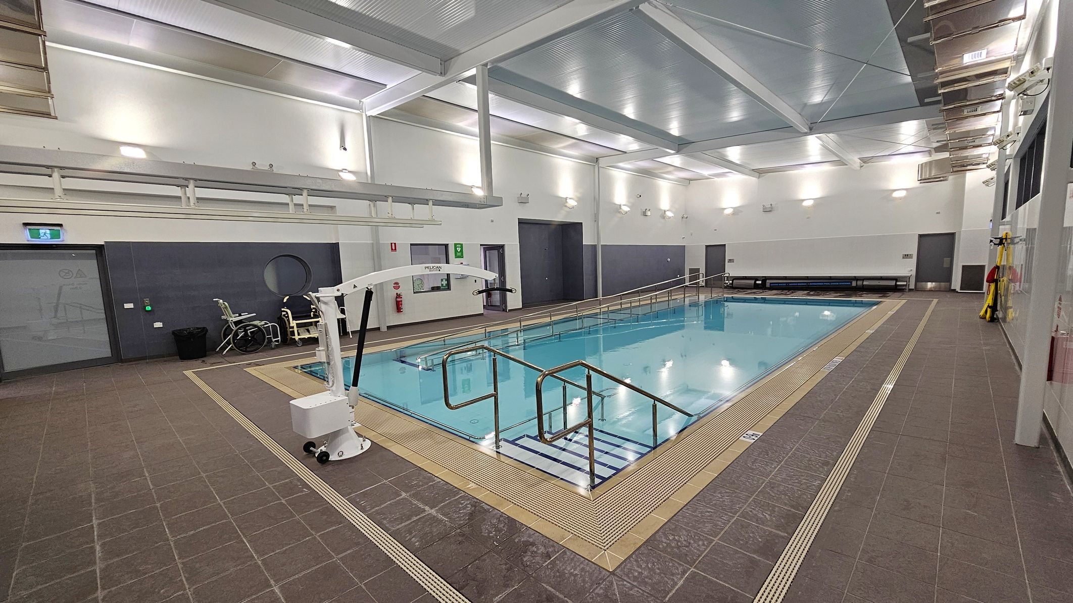 Indoor swimming pool with a tiled deck surrounding it. On the left, there's a pool lift and a wheelchair near a grey door with a circular window. The pool has metal handrails and a ramp for accessibility. The walls are white with several lights, and there are benches along the far wall. The ceiling is high with exposed beams and bright lighting. The floor is brown tile with drainage grates around the pool's edge. Indoor swimming pool with a tiled deck surrounding it. On the left, there's a pool lift and a wheelchair near a grey door with a circular window. The pool has metal handrails and a ramp for accessibility. The walls are white with several lights, and there are benches along the far wall. The ceiling is high with exposed beams and bright lighting. The floor is brown tile with drainage grates around the pool's edge.