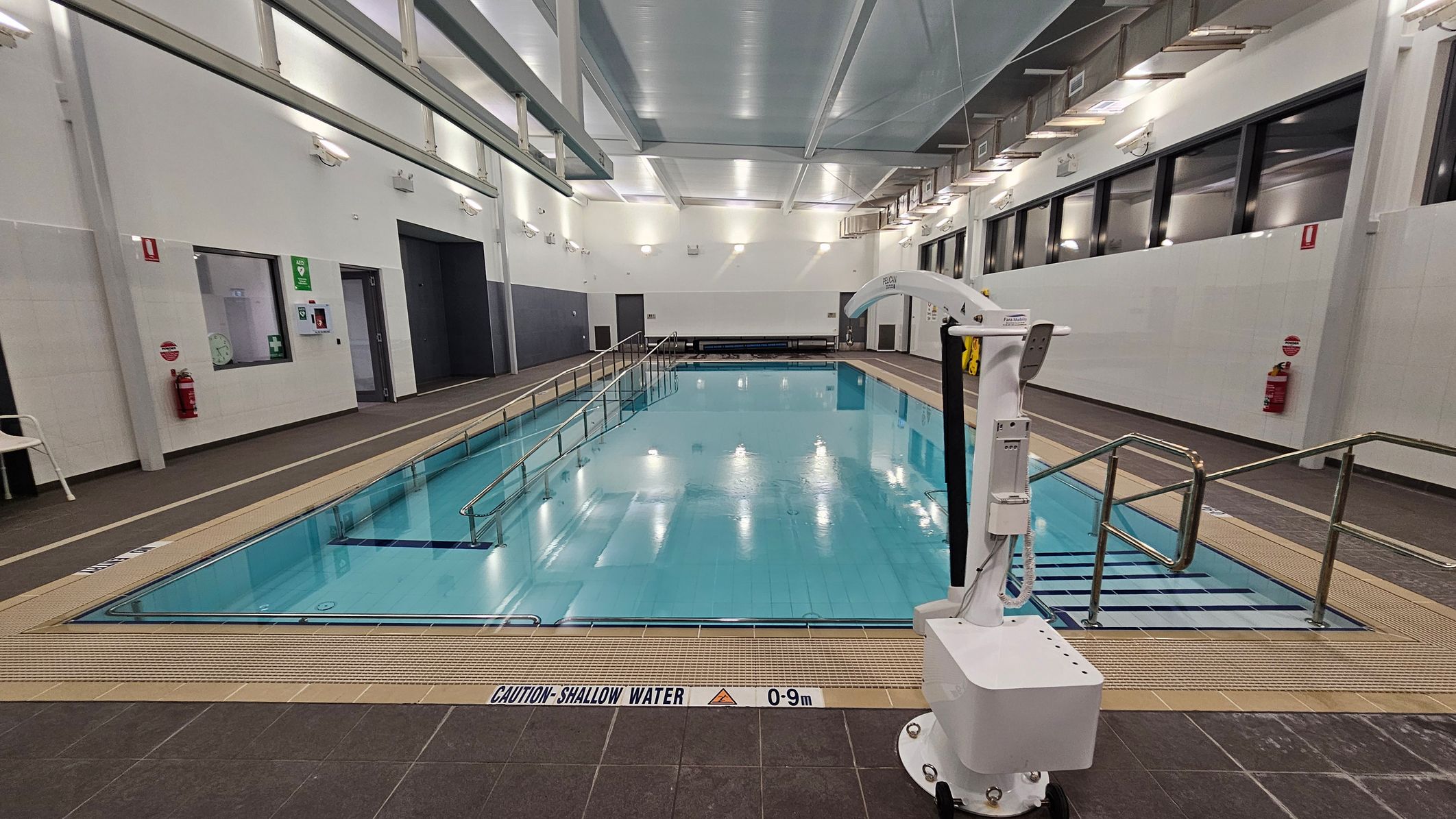 An indoor swimming pool with clear blue water is in the centre. On the right, a white pool lift is positioned near metal handrails leading into the pool. The left side features a wall with emergency equipment, including a fire extinguisher and a first aid kit. The ceiling is high with exposed beams and lights. The floor surrounding the pool is tiled in dark brown, with a caution sign indicating shallow water. Large windows line the right wall. An indoor swimming pool with clear blue water is in the centre. On the right, a white pool lift is positioned near metal handrails leading into the pool. The left side features a wall with emergency equipment, including a fire extinguisher and a first aid kit. The ceiling is high with exposed beams and lights. The floor surrounding the pool is tiled in dark brown, with a caution sign indicating shallow water. Large windows line the right wall.