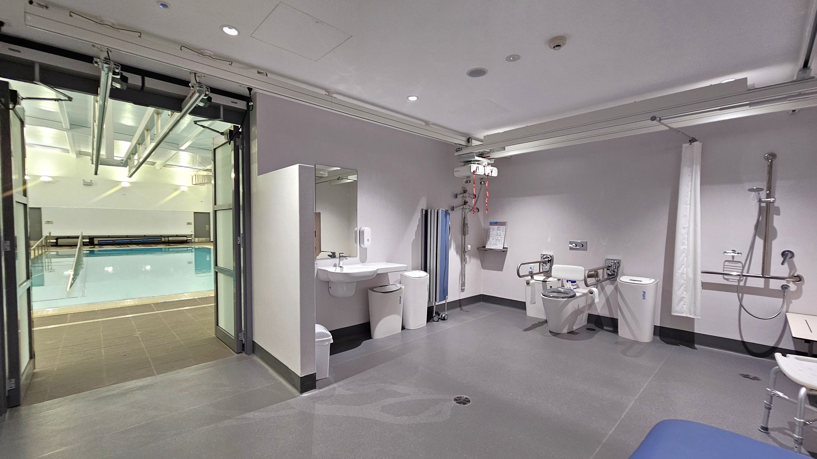 On the left, there is a swimming pool visible through large glass doors. The pool area is well-lit with overhead lights. In the middle, a bathroom area features a sink with a mirror, soap dispenser, and bins underneath. On the right, there is a toilet with support rails, a shower with a handheld nozzle, and a shower chair. The ceiling has recessed lighting and a track system for a hoist. The floor is smooth and grey. On the left, there is a swimming pool visible through large glass doors. The pool area is well-lit with overhead lights. In the middle, a bathroom area features a sink with a mirror, soap dispenser, and bins underneath. On the right, there is a toilet with support rails, a shower with a handheld nozzle, and a shower chair. The ceiling has recessed lighting and a track system for a hoist. The floor is smooth and grey.