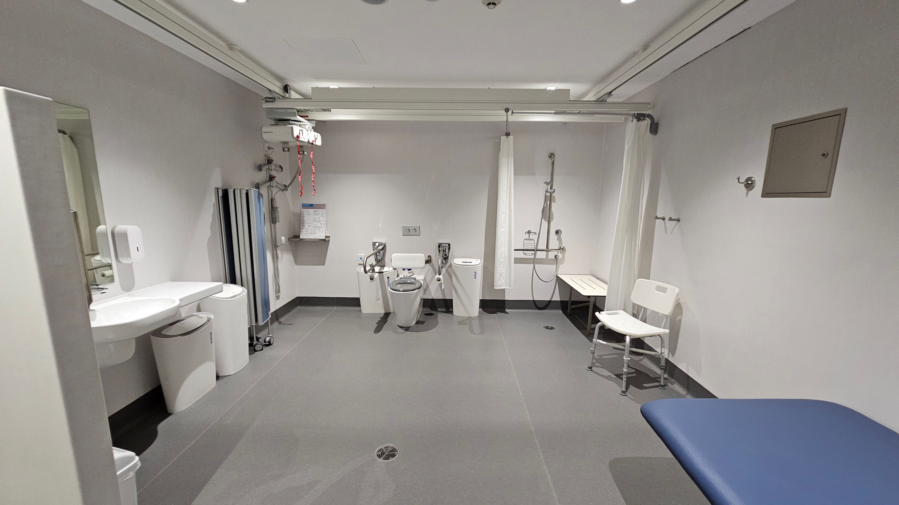 A spacious, accessible bathroom with grey tiled flooring and a white ceiling. On the left, there is a sink with a soap dispenser and bins underneath. In the centre, a toilet with support rails is visible, along with a wall-mounted hoist system. To the right, a shower area with a curtain, a bench, and a white chair is present. A blue padded bench is in the foreground on the right. The walls are plain with a small notice board on the right. A spacious, accessible bathroom with grey tiled flooring and a white ceiling. On the left, there is a sink with a soap dispenser and bins underneath. In the centre, a toilet with support rails is visible, along with a wall-mounted hoist system. To the right, a shower area with a curtain, a bench, and a white chair is present. A blue padded bench is in the foreground on the right. The walls are plain with a small notice board on the right.