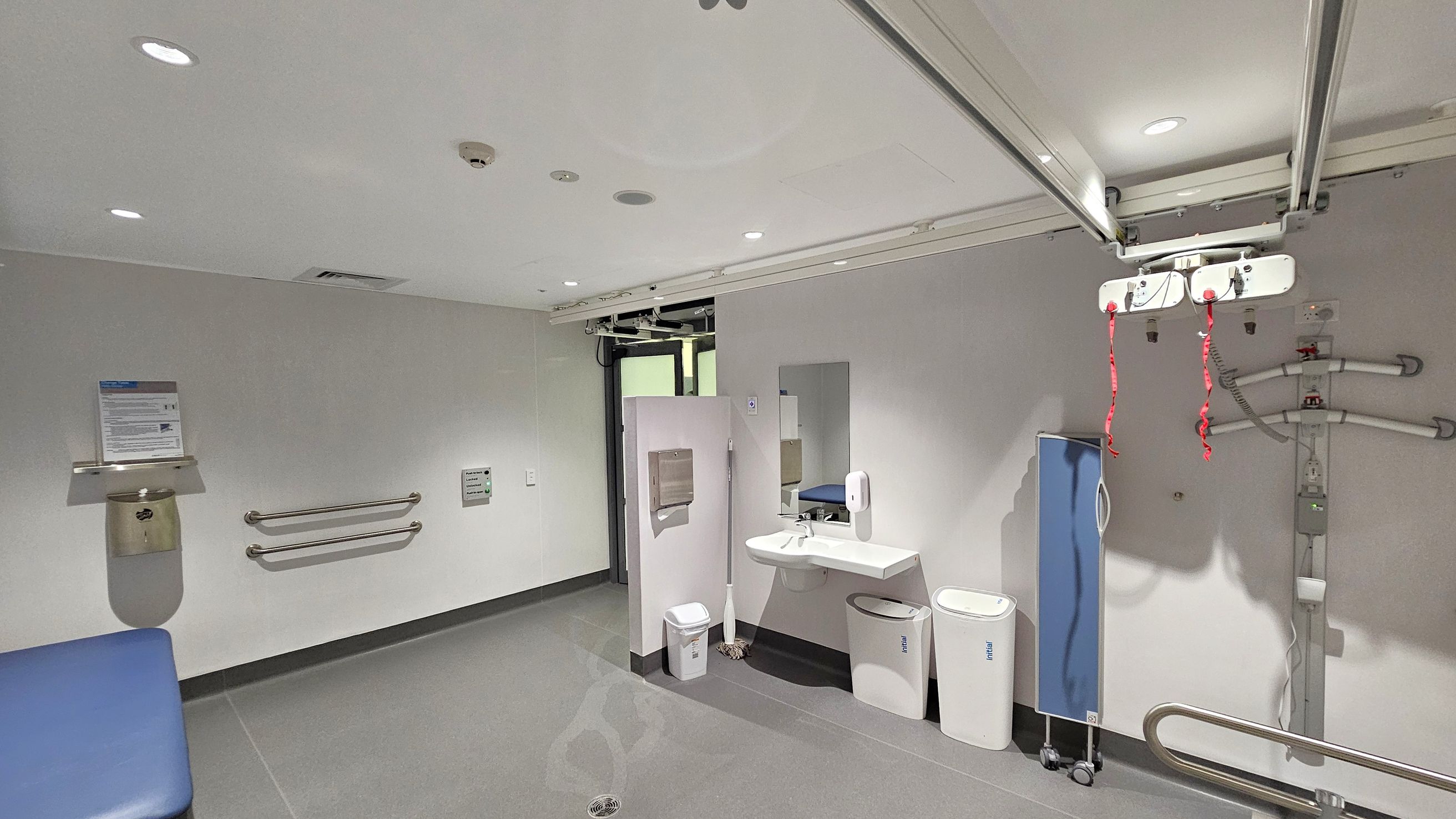 A hospital room with a grey floor and white ceiling. On the left, there are two horizontal grab bars and a wall-mounted bin with a sign above it. In the centre, a sink with a mirror, soap dispenser, and two bins below. A mop is leaning against the wall. On the right, a blue privacy screen on wheels and a ceiling-mounted hoist with red straps. The room has recessed lighting and a door in the background. A hospital room with a grey floor and white ceiling. On the left, there are two horizontal grab bars and a wall-mounted bin with a sign above it. In the centre, a sink with a mirror, soap dispenser, and two bins below. A mop is leaning against the wall. On the right, a blue privacy screen on wheels and a ceiling-mounted hoist with red straps. The room has recessed lighting and a door in the background.