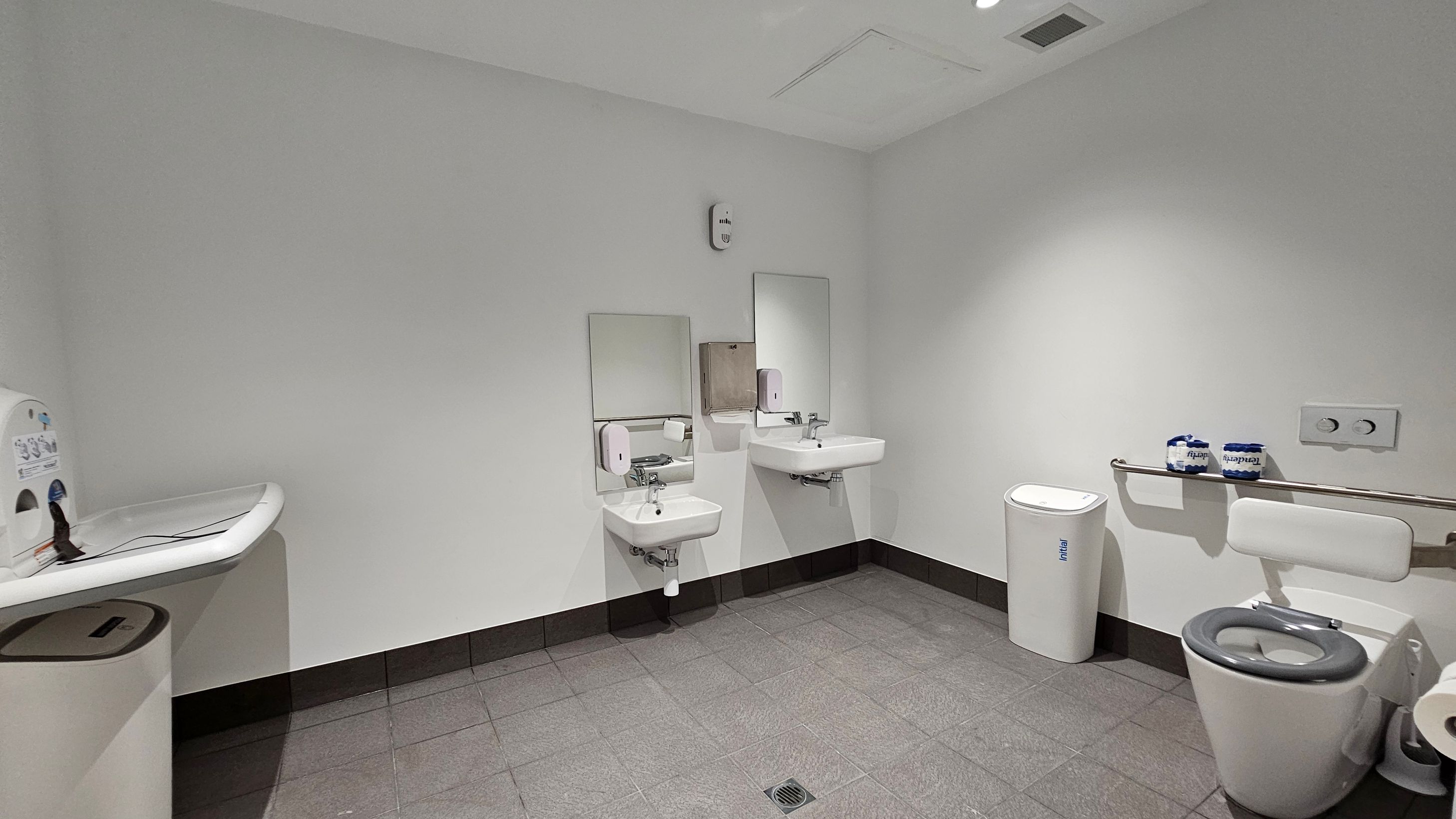 A public restroom with grey tiled flooring and white walls. On the left, a baby changing station is mounted above a bin. In the centre, two wall-mounted sinks with mirrors and soap dispensers are visible. On the right, a toilet with a support bar and two rolls of toilet paper on top. A sanitary bin is placed beside the toilet. The ceiling is plain with recessed lighting. A public restroom with grey tiled flooring and white walls. On the left, a baby changing station is mounted above a bin. In the centre, two wall-mounted sinks with mirrors and soap dispensers are visible. On the right, a toilet with a support bar and two rolls of toilet paper on top. A sanitary bin is placed beside the toilet. The ceiling is plain with recessed lighting.