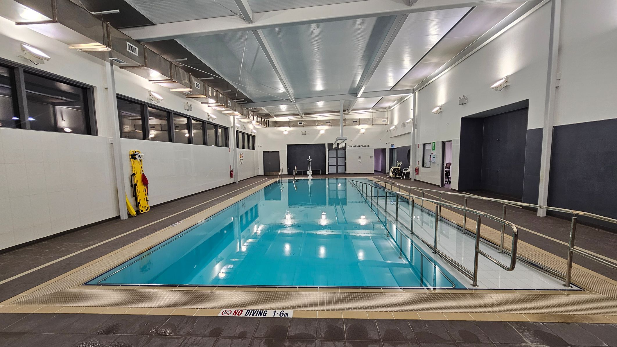 An indoor swimming pool with clear blue water is in the centre. On the left, there are large windows and a yellow rescue board against the wall. On the right, there is a ramp with metal railings leading into the pool. The walls are white with several lights, and there are doors labelled "Changing Places" in the background. The ceiling is high with exposed beams and ventilation ducts. The floor around the pool is tiled in a dark colour. An indoor swimming pool with clear blue water is in the centre. On the left, there are large windows and a yellow rescue board against the wall. On the right, there is a ramp with metal railings leading into the pool. The walls are white with several lights, and there are doors labelled "Changing Places" in the background. The ceiling is high with exposed beams and ventilation ducts. The floor around the pool is tiled in a dark colour.