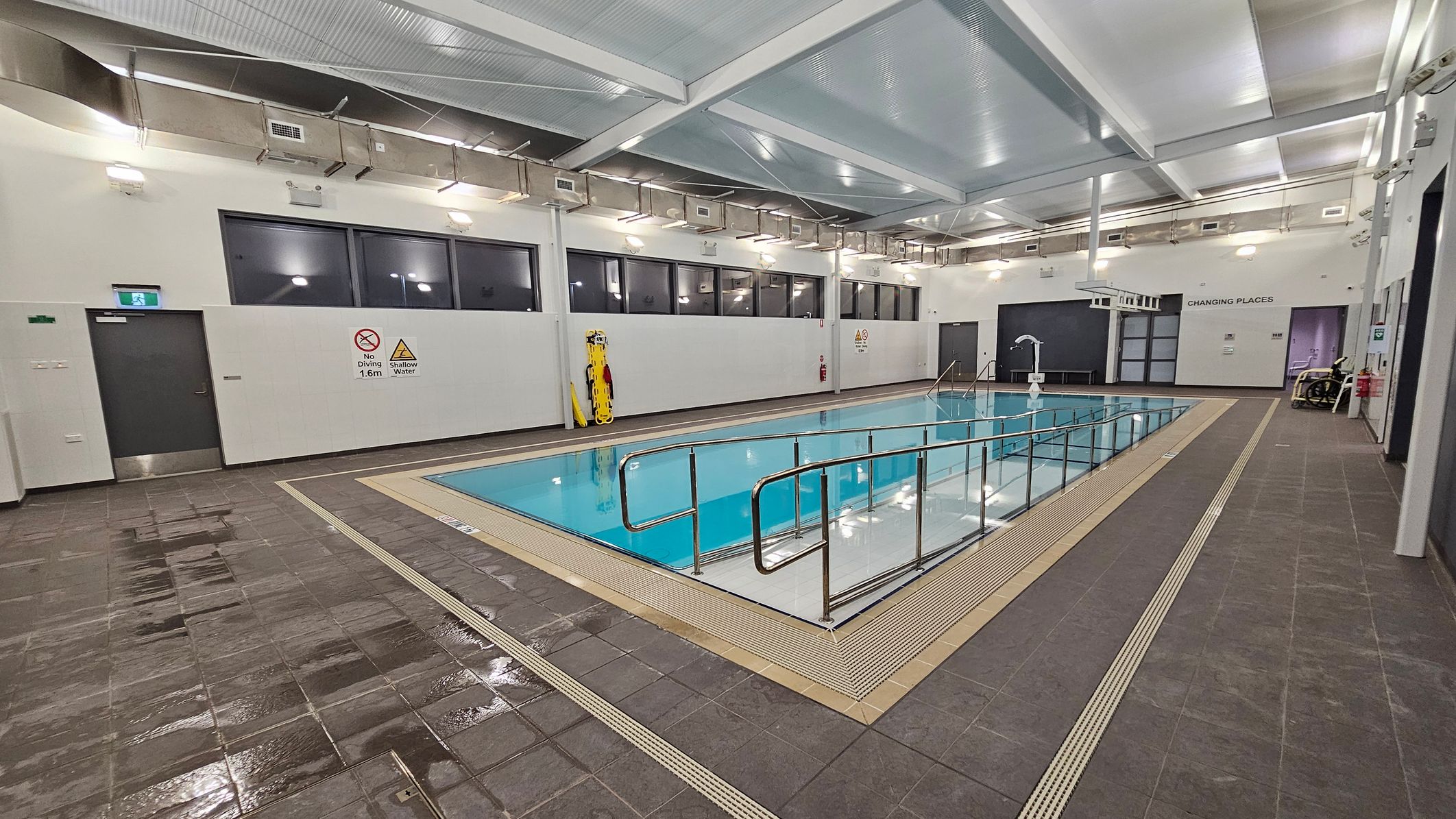 An indoor swimming pool with a metal handrail along its edge. The pool is surrounded by wet, brown tiles. On the left wall, there are signs indicating "No Diving" and "Shallow Water," along with a door and a mounted rescue board. The right side features a door labelled "Changing Places" and a wheelchair. The ceiling is high with exposed beams and lighting fixtures. Large windows line the upper part of the walls. An indoor swimming pool with a metal handrail along its edge. The pool is surrounded by wet, brown tiles. On the left wall, there are signs indicating "No Diving" and "Shallow Water," along with a door and a mounted rescue board. The right side features a door labelled "Changing Places" and a wheelchair. The ceiling is high with exposed beams and lighting fixtures. Large windows line the upper part of the walls.