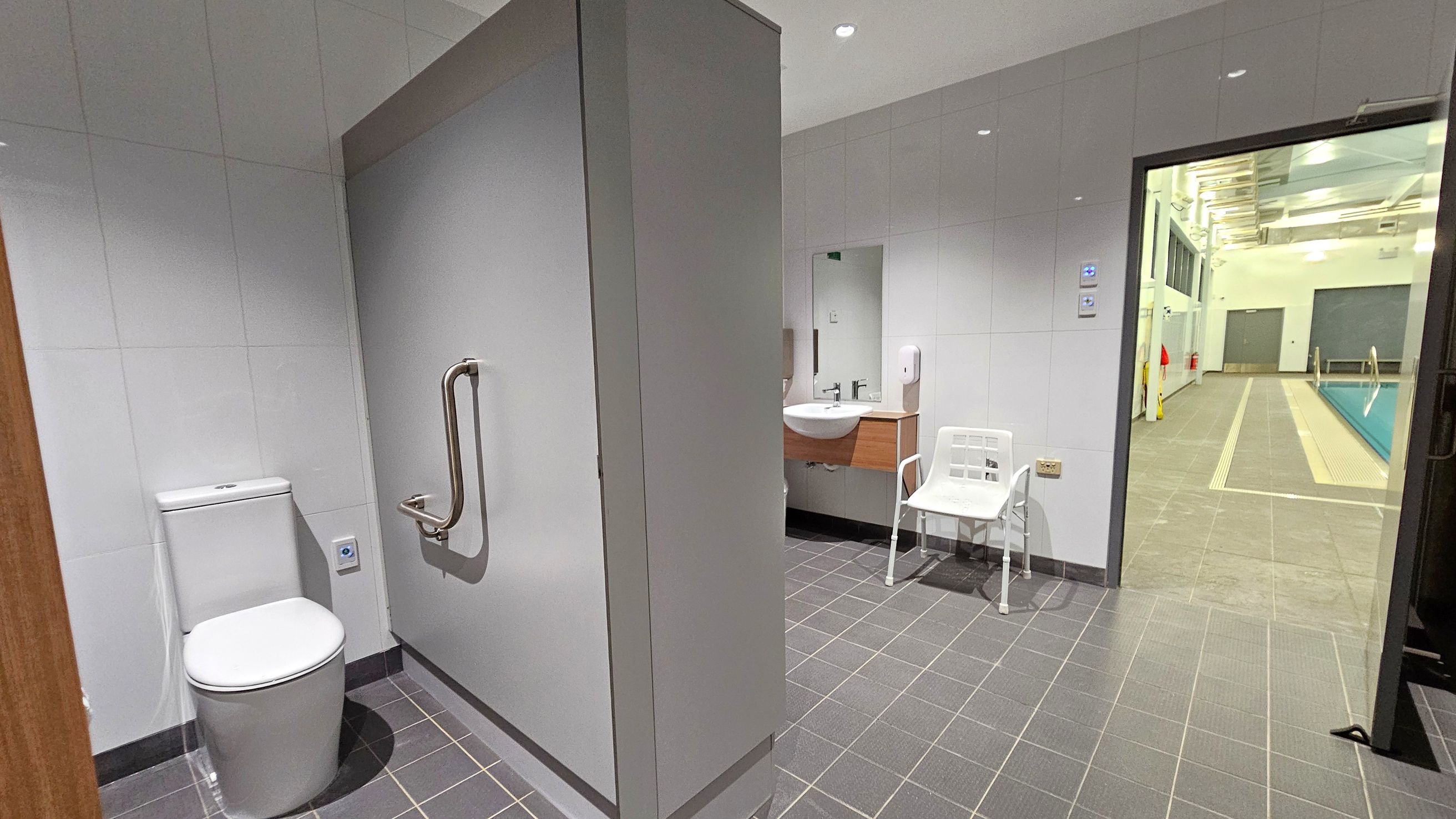 A bathroom with grey tiled flooring and white tiled walls. On the left, there is a toilet with a metal grab bar. In the centre, a sink with a mirror above it and a white chair nearby. On the right, an open door leads to a swimming pool area with a tiled walkway and a pool visible. The ceiling has recessed lighting. A bathroom with grey tiled flooring and white tiled walls. On the left, there is a toilet with a metal grab bar. In the centre, a sink with a mirror above it and a white chair nearby. On the right, an open door leads to a swimming pool area with a tiled walkway and a pool visible. The ceiling has recessed lighting.
