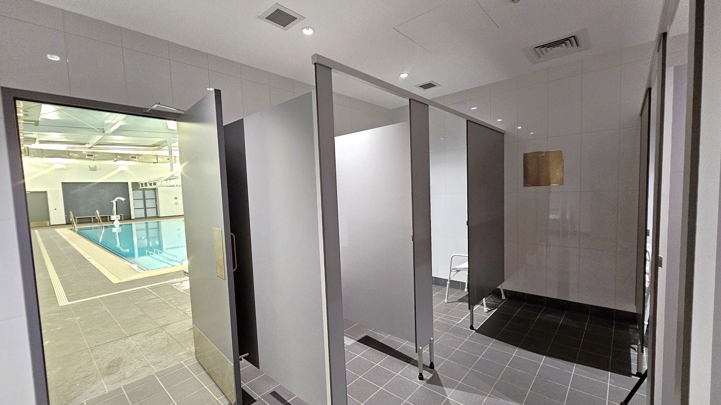 On the left, there is a view of an indoor swimming pool with a tiled deck and metal handrails. The pool area is well-lit with overhead lights and has a high ceiling. On the right, there are changing cubicles with grey doors and tiled flooring. A small white chair is visible inside one of the cubicles. The ceiling is white with recessed lighting and ventilation panels. On the left, there is a view of an indoor swimming pool with a tiled deck and metal handrails. The pool area is well-lit with overhead lights and has a high ceiling. On the right, there are changing cubicles with grey doors and tiled flooring. A small white chair is visible inside one of the cubicles. The ceiling is white with recessed lighting and ventilation panels.