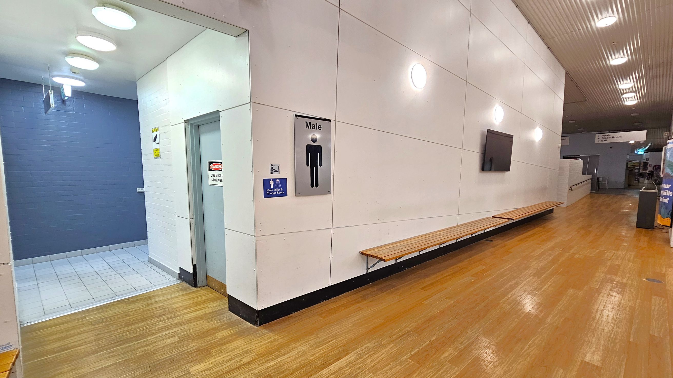 A corridor with wooden flooring and a white panelled wall on the right. A long wooden bench is attached to the wall. A sign indicating a male change room is visible. On the left, a tiled area leads to a door marked "Danger Chemical Storage." The ceiling has circular lights, and further down the corridor, there are signs and a vending machine. A corridor with wooden flooring and a white panelled wall on the right. A long wooden bench is attached to the wall. A sign indicating a male change room is visible. On the left, a tiled area leads to a door marked "Danger Chemical Storage." The ceiling has circular lights, and further down the corridor, there are signs and a vending machine.