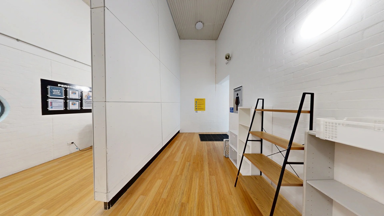 A room with light wooden flooring and white walls. On the right, there are two sets of shelves, one with a black frame and wooden shelves, the other white. In the centre, a black mat is on the floor near a door with a sign above it. On the left wall, a black-framed bulletin board displays various notices. The ceiling is light green with a circular light fixture. A room with light wooden flooring and white walls. On the right, there are two sets of shelves, one with a black frame and wooden shelves, the other white. In the centre, a black mat is on the floor near a door with a sign above it. On the left wall, a black-framed bulletin board displays various notices. The ceiling is light green with a circular light fixture.