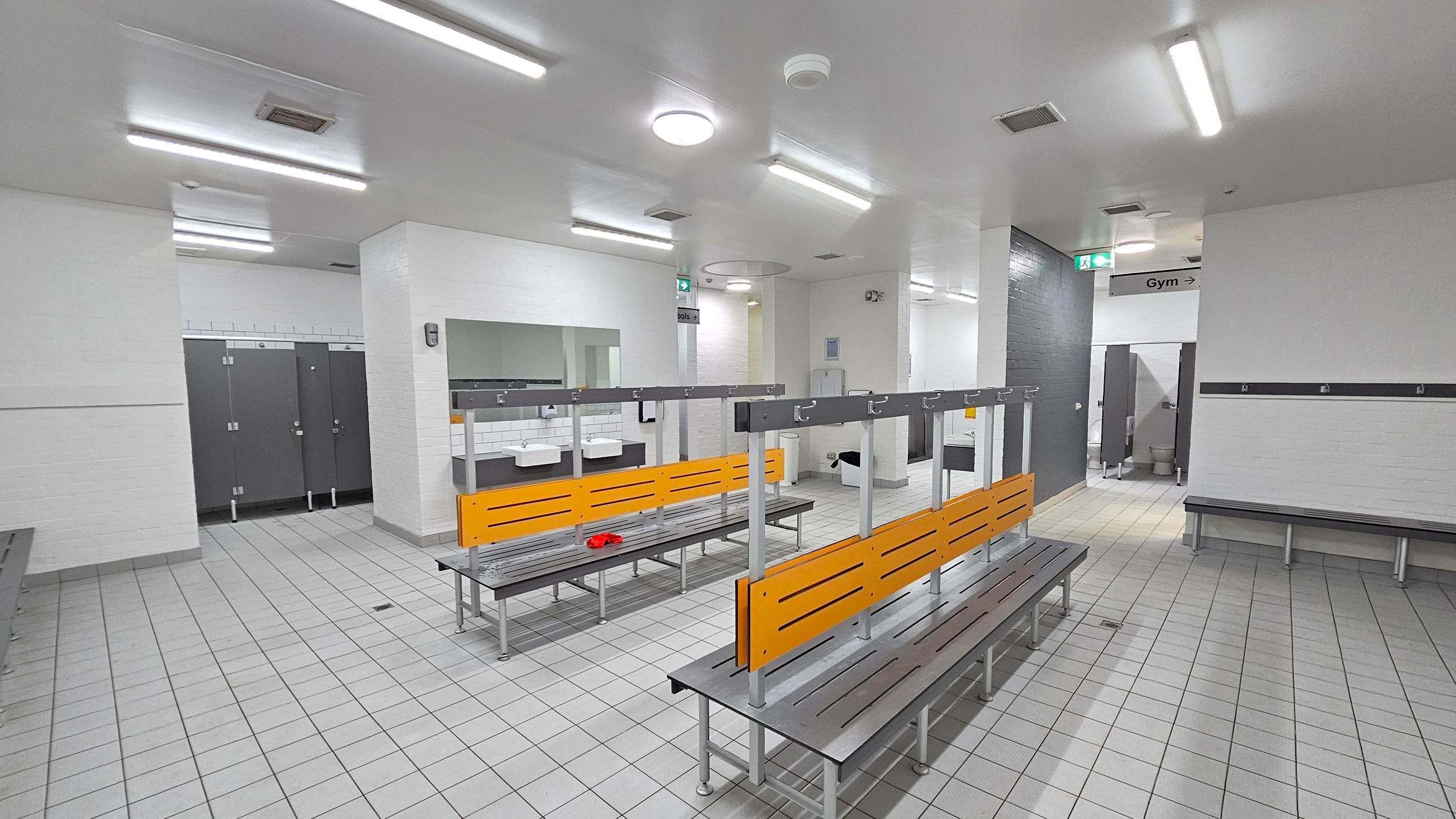 A spacious locker room with grey tiled flooring and white walls. In the centre, there are benches with orange backrests and metal hooks above. To the left, there are several closed grey lockers. In the background, sinks with mirrors are visible. On the right, a sign indicates the direction to the gym. The ceiling is white with fluorescent lights. A red item is placed on one of the benches. A spacious locker room with grey tiled flooring and white walls. In the centre, there are benches with orange backrests and metal hooks above. To the left, there are several closed grey lockers. In the background, sinks with mirrors are visible. On the right, a sign indicates the direction to the gym. The ceiling is white with fluorescent lights. A red item is placed on one of the benches.