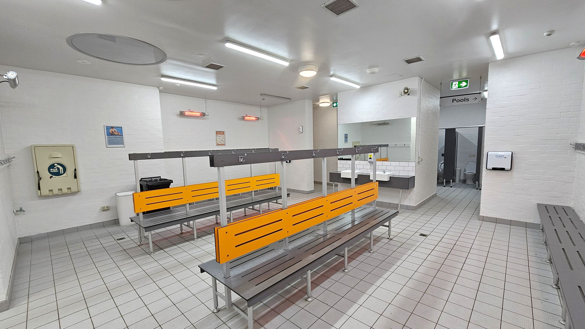 A changing room with grey tiled flooring and a white ceiling with fluorescent lights. In the centre, there are benches with orange backrests and metal frames. On the left wall, there is a baby changing station and a waste bin. The right side has a sign indicating the direction to the pools and a glimpse of a toilet cubicle. Sinks with mirrors are visible near the entrance to the toilets. The walls are white brick, and there are coat hooks along the left wall. A changing room with grey tiled flooring and a white ceiling with fluorescent lights. In the centre, there are benches with orange backrests and metal frames. On the left wall, there is a baby changing station and a waste bin. The right side has a sign indicating the direction to the pools and a glimpse of a toilet cubicle. Sinks with mirrors are visible near the entrance to the toilets. The walls are white brick, and there are coat hooks along the left wall.