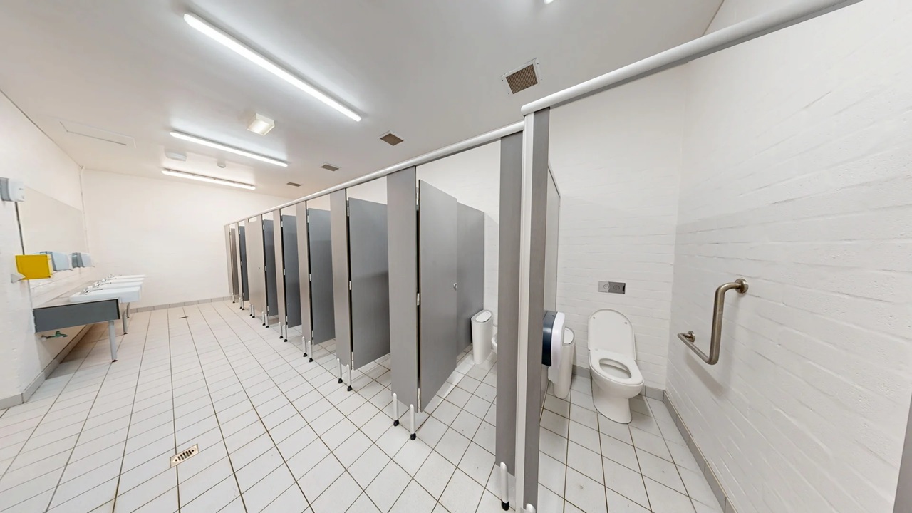 A public restroom with a row of toilet cubicles on the right, each with grey doors and partitions. The first cubicle door is open, revealing a white toilet and a grab bar on the wall. On the left, there are sinks with a yellow soap dispenser and a hand dryer. The floor is covered with white tiles arranged diagonally, and the ceiling has fluorescent lights. The walls are plain white, contributing to a clean and minimalistic appearance. A public restroom with a row of toilet cubicles on the right, each with grey doors and partitions. The first cubicle door is open, revealing a white toilet and a grab bar on the wall. On the left, there are sinks with a yellow soap dispenser and a hand dryer. The floor is covered with white tiles arranged diagonally, and the ceiling has fluorescent lights. The walls are plain white, contributing to a clean and minimalistic appearance.