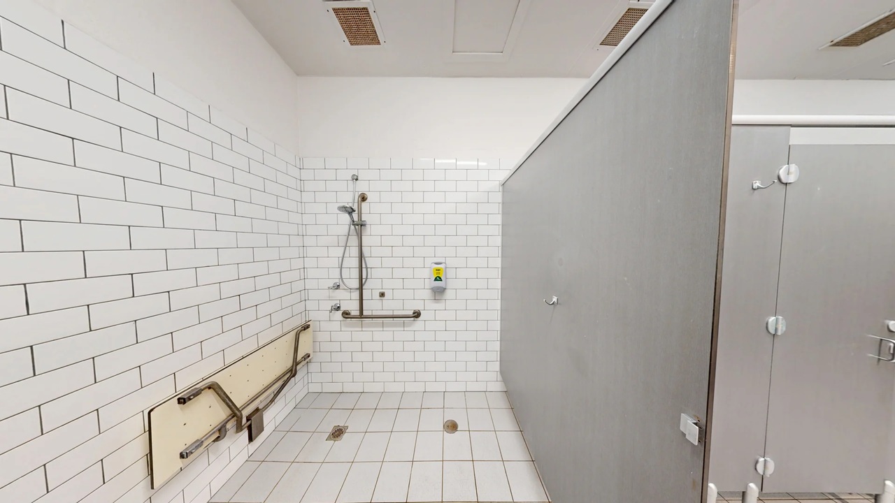 A bathroom with white subway tiles on the walls and floor. On the left, there is a foldable bench with metal support bars. In the centre, a showerhead is mounted on the wall with a grab bar below it. A soap dispenser is also visible. On the right, a grey partition separates the shower area from another section. The ceiling is plain white with ventilation panels. A bathroom with white subway tiles on the walls and floor. On the left, there is a foldable bench with metal support bars. In the centre, a showerhead is mounted on the wall with a grab bar below it. A soap dispenser is also visible. On the right, a grey partition separates the shower area from another section. The ceiling is plain white with ventilation panels.