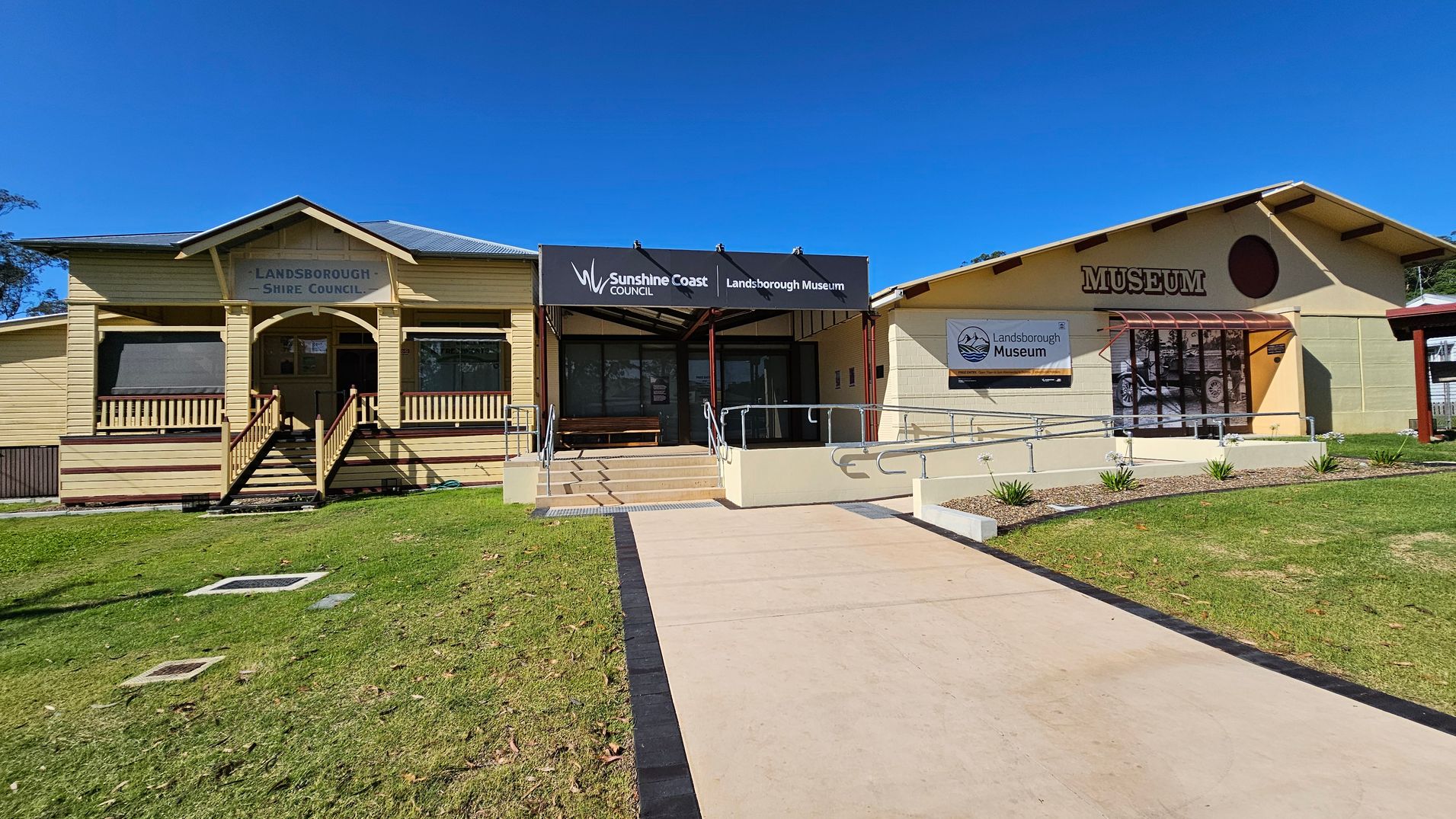 A building with two sections. On the left, a yellow wooden structure with a sign reading "Landsborough Shire Council" above a small porch with steps. In the centre, a covered entrance with a sign for "Sunshine Coast Council Landsborough Museum" and a ramp leading up. On the right, a beige building with a large "MUSEUM" sign and a window displaying a "Landsborough Museum" banner. The foreground features a concrete path and a grassy area with small plants. A building with two sections. On the left, a yellow wooden structure with a sign reading "Landsborough Shire Council" above a small porch with steps. In the centre, a covered entrance with a sign for "Sunshine Coast Council Landsborough Museum" and a ramp leading up. On the right, a beige building with a large "MUSEUM" sign and a window displaying a "Landsborough Museum" banner. The foreground features a concrete path and a grassy area with small plants.