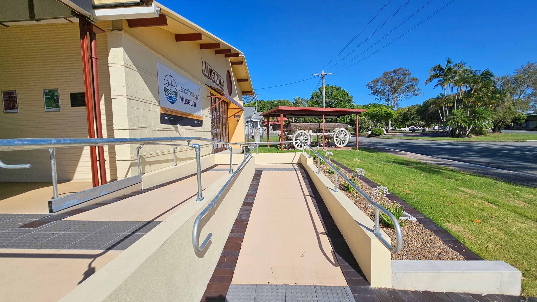 A museum entrance with a ramp and metal railings on the left. The building is light-coloured with a sign reading "Landsborough Museum." Posters are displayed on the wall. On the right, a grassy area with a covered wagon under a red shelter. Trees and a road are visible in the background. The sky is clear and blue. A museum entrance with a ramp and metal railings on the left. The building is light-coloured with a sign reading "Landsborough Museum." Posters are displayed on the wall. On the right, a grassy area with a covered wagon under a red shelter. Trees and a road are visible in the background. The sky is clear and blue.